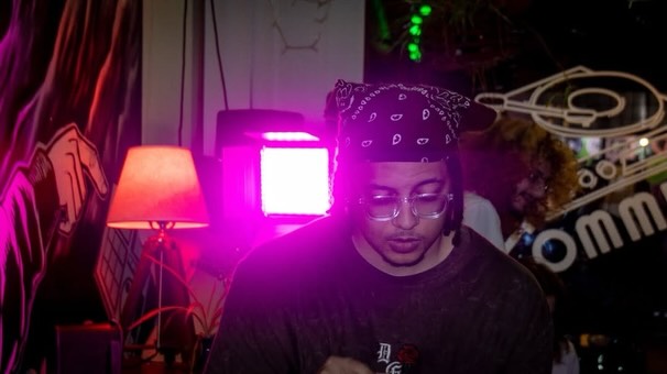 Person wearing a bandana and glasses in a dimly lit room with colorful lights and a lamp.
