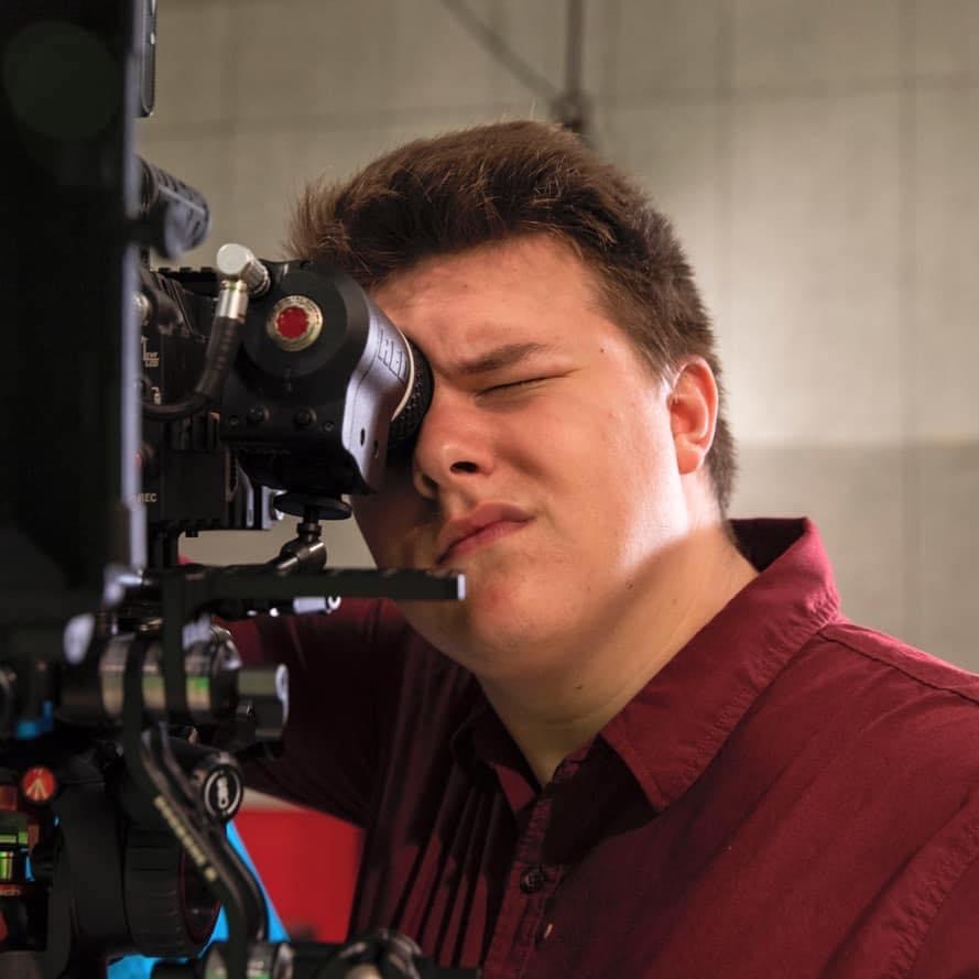 Young man with closed eyes looking into camera lens, wearing a maroon shirt, in a studio setting.