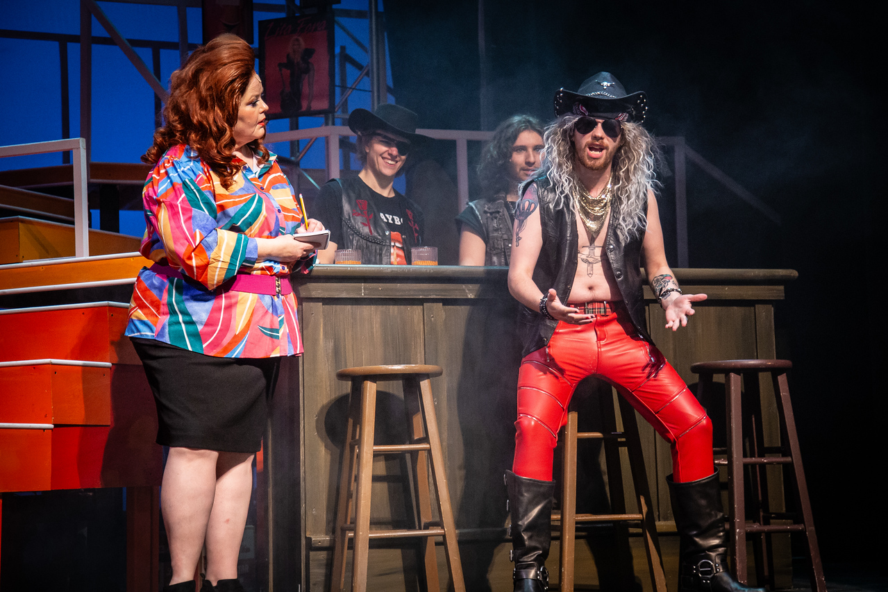 Group of people on stage, one person in colorful jacket and others in costumes, some smiling and talking.