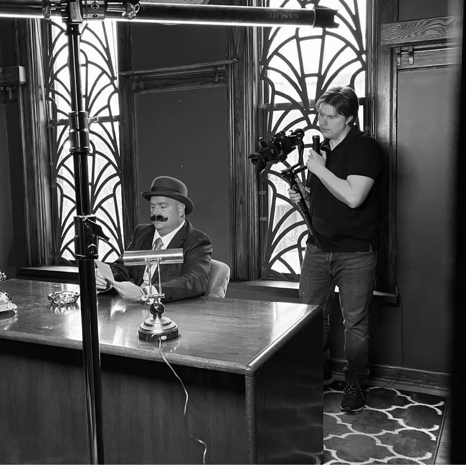 Person with a mustache and hat sitting at a desk, another person standing with a camera, in a room with decorative windows.