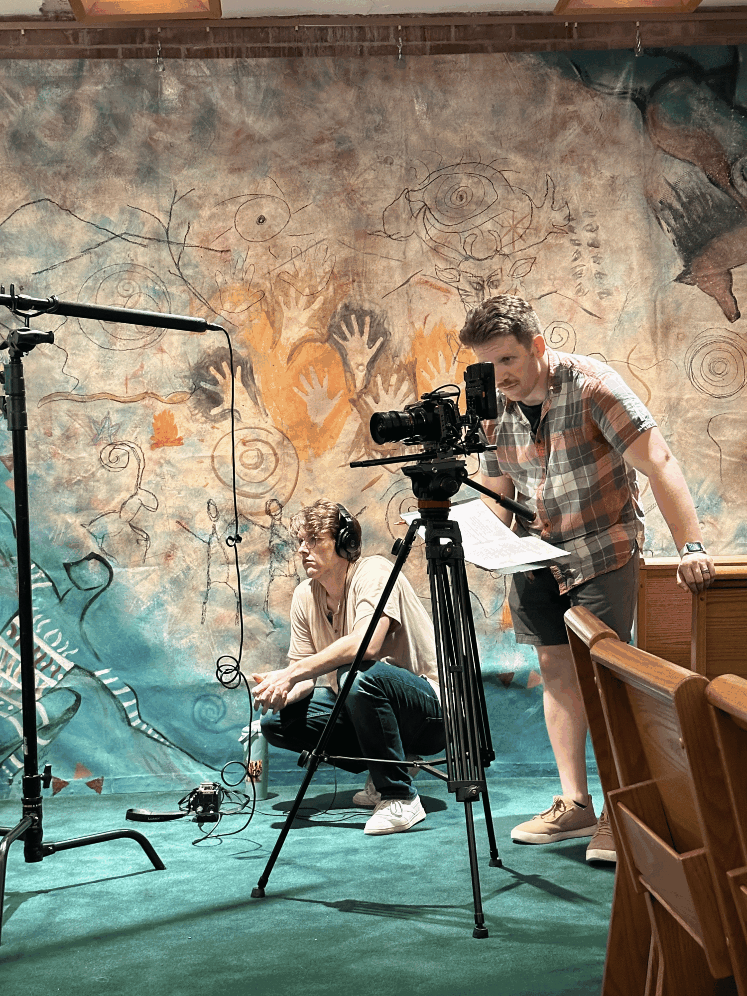 Two people setting up camera equipment in front of a colorful, abstract mural background in a room with wooden chairs.