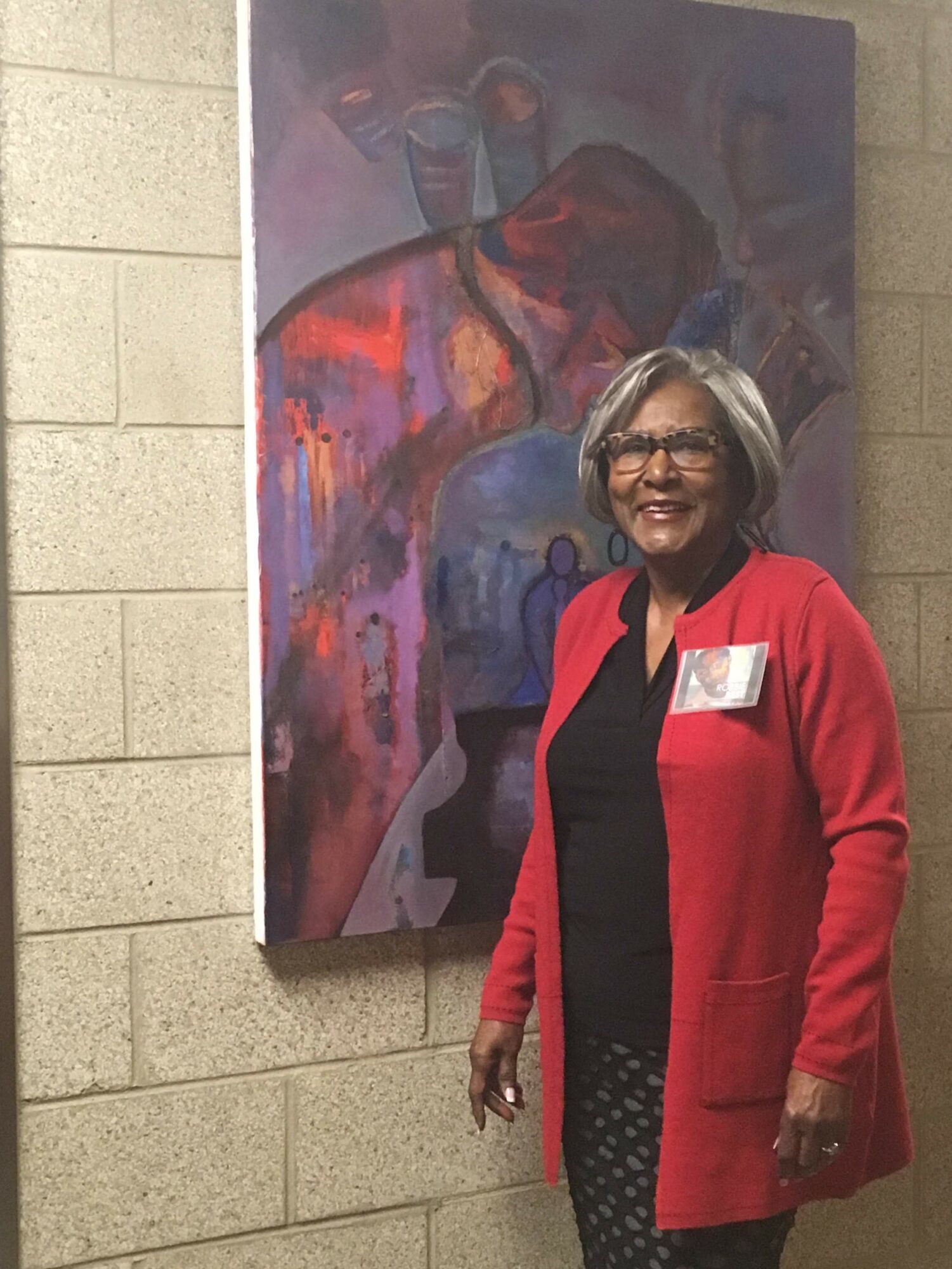 Smiling woman with glasses and gray hair stands in front of colorful abstract painting on beige brick wall, wearing a red coat and black dress.