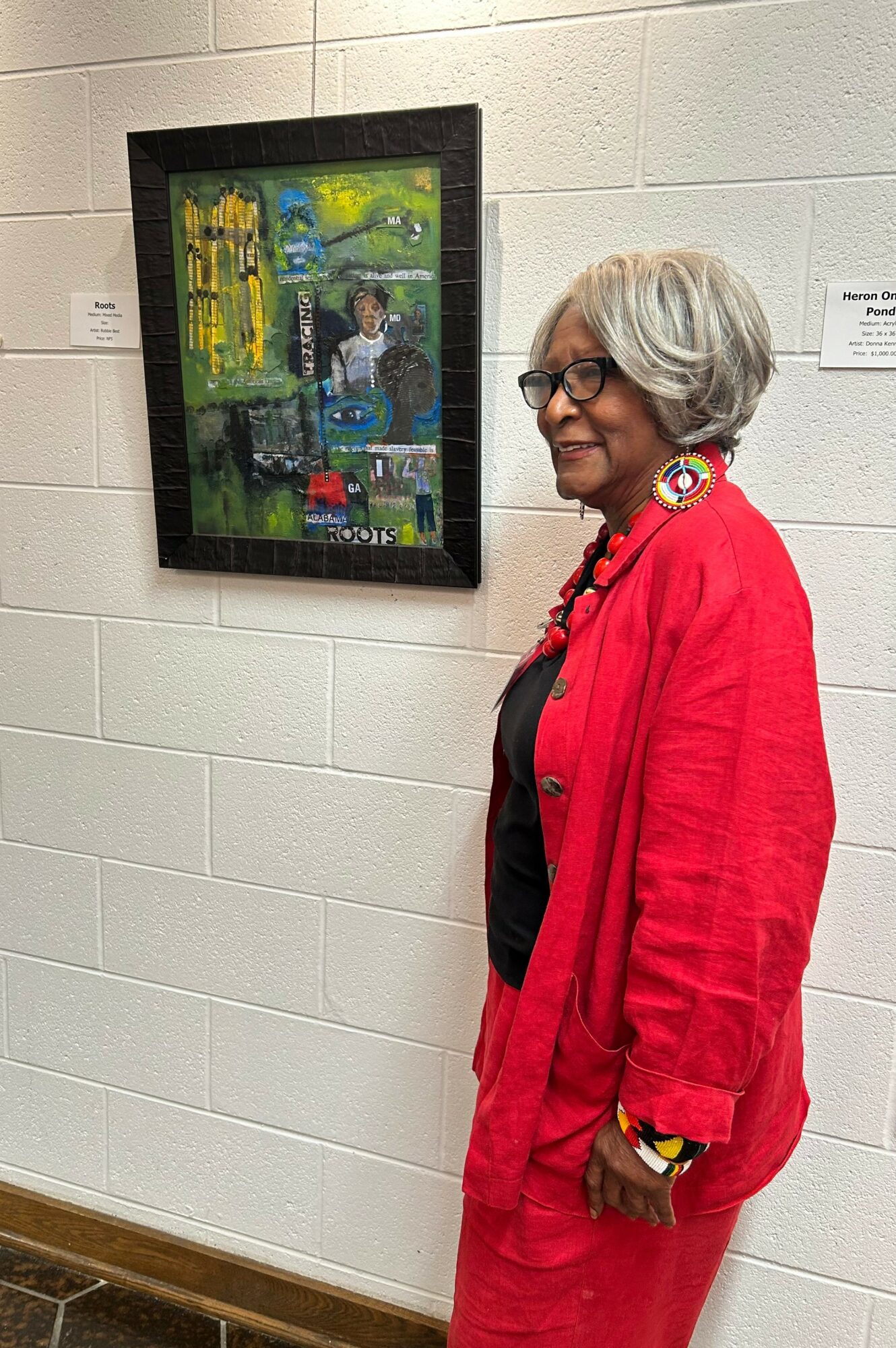 Woman with gray hair and glasses wearing a red jacket, standing next to a colorful framed painting on a white wall.