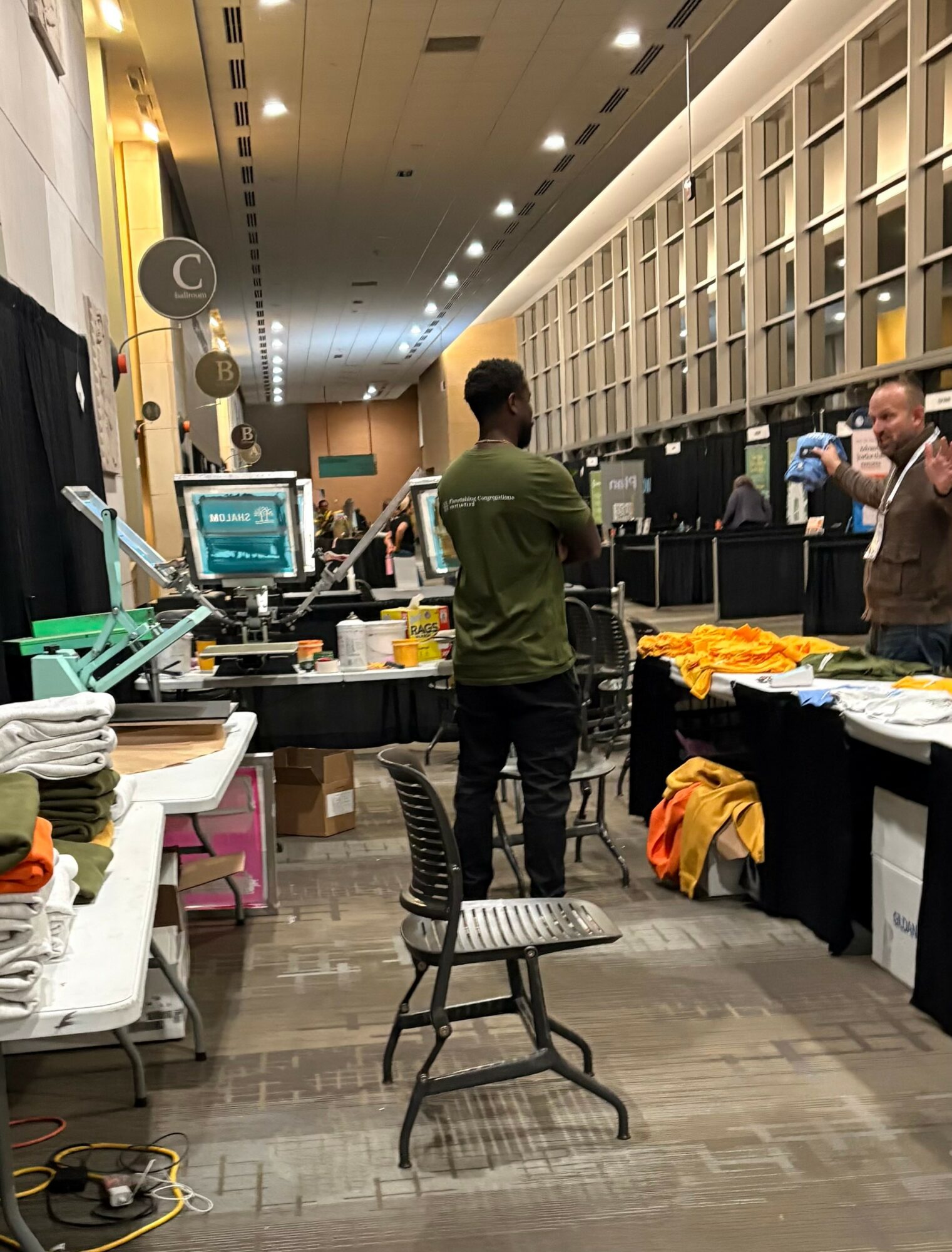 People working at a computer station in a large room with shelves, tables, and equipment, viewed from the front.