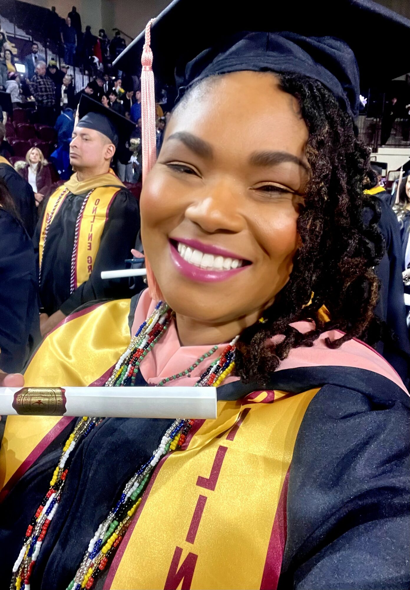 Smiling woman in graduation cap and gown at ceremony, other graduates in background, colorful cords and medals, indoor setting.