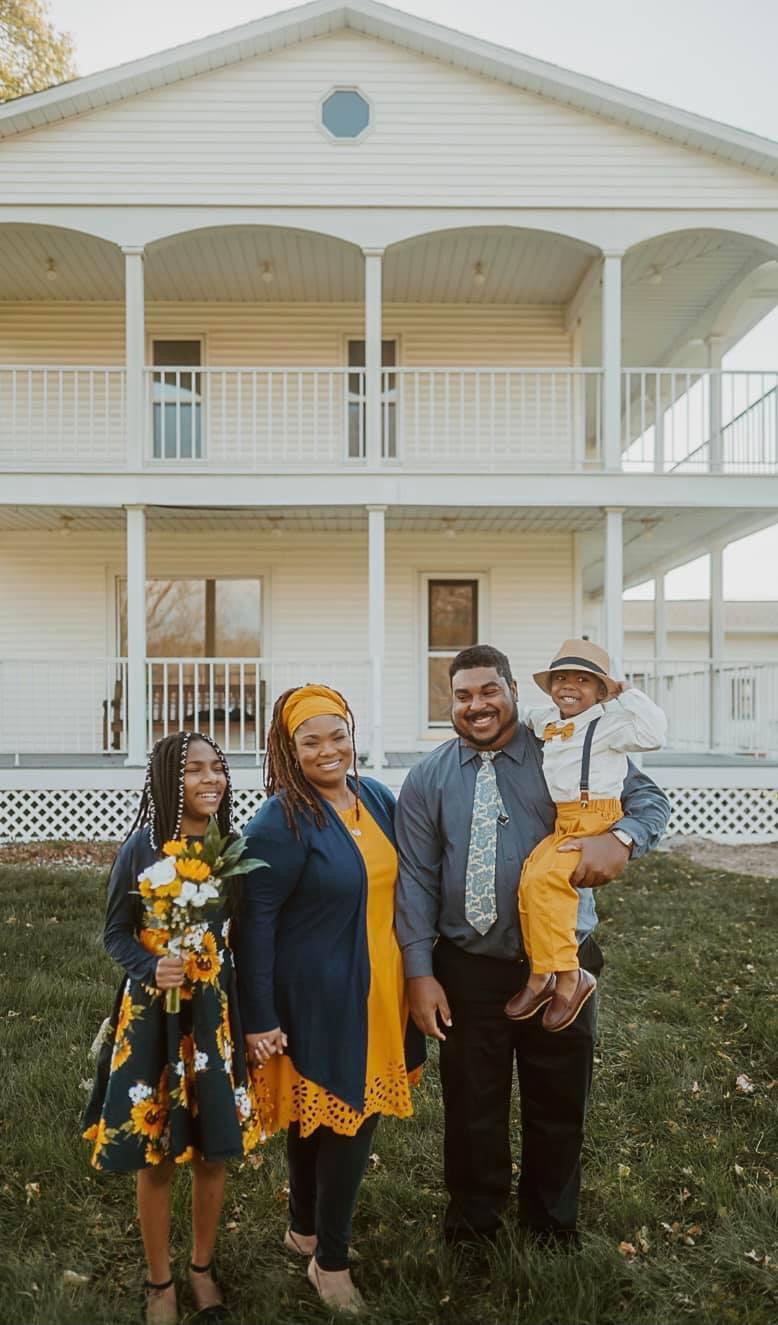 Family of four standing in front of a large white house with a porch, smiling and posing for the photo.