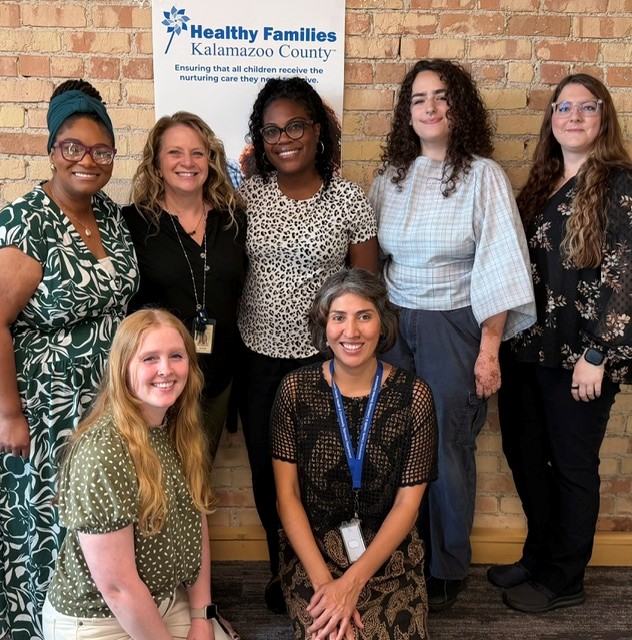 Group of seven women standing in front of a brick wall and a sign for Healthy Families Kalamazoo County, smiling.