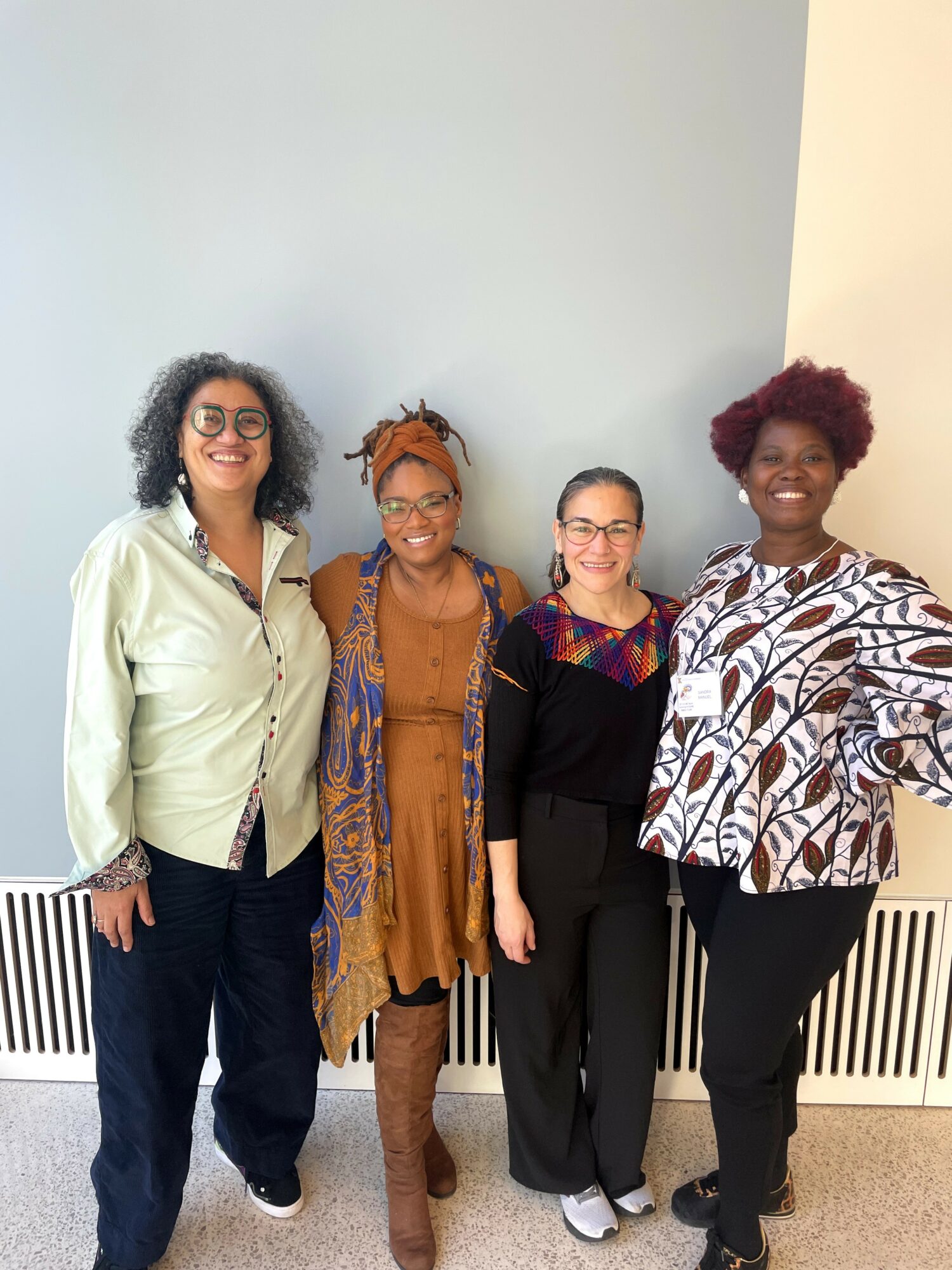 Four women standing together indoors, smiling, against a light-colored wall with a radiator below.