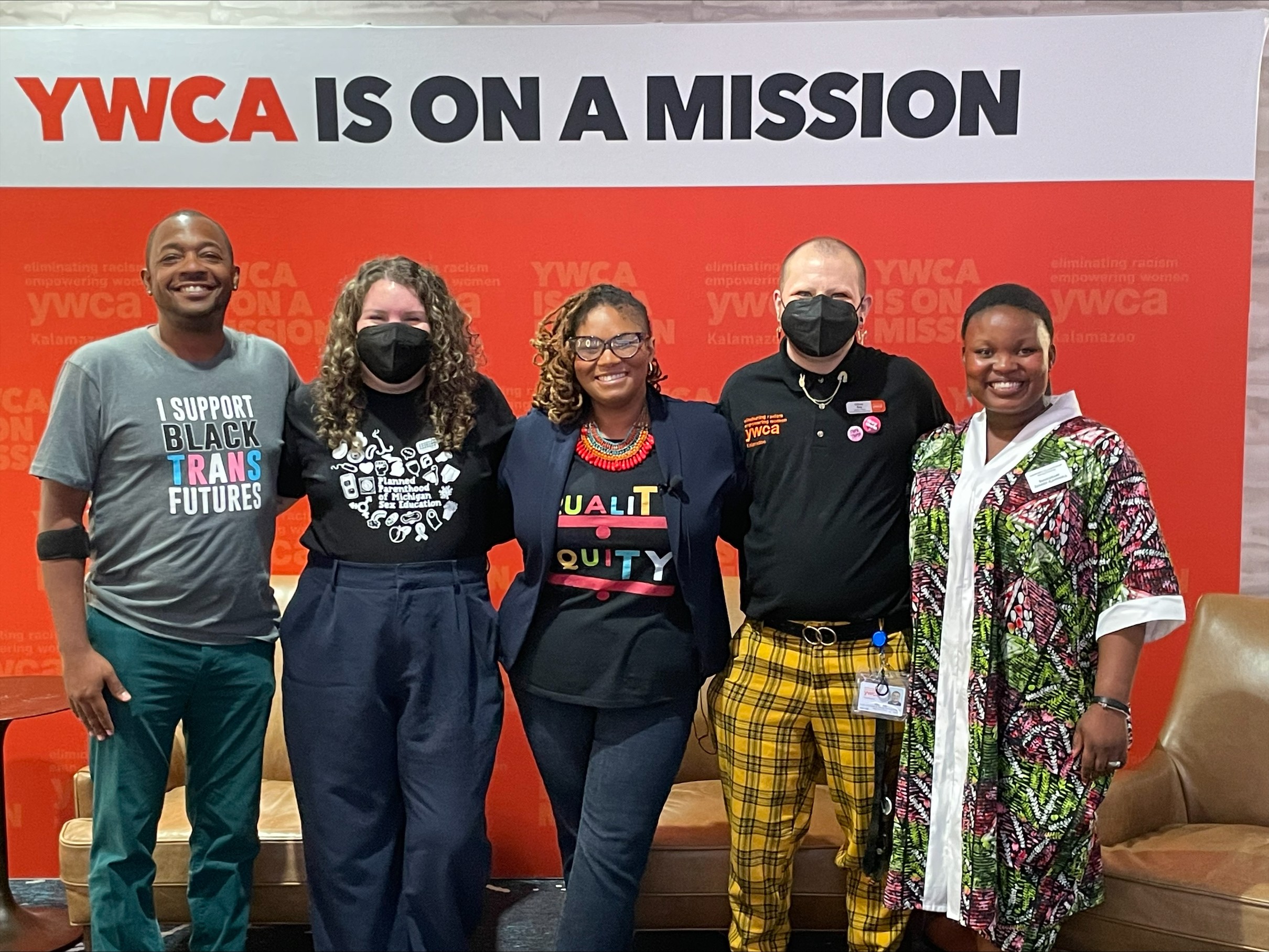 Group of five people standing in front of a YWCA banner, smiling, wearing casual and colorful clothing, some with masks.
