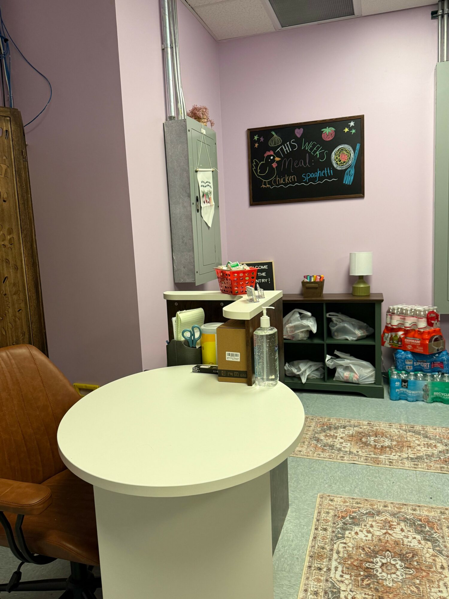 A round white table with a brown chair, a shelf with cups and bottles, and a pink wall with a chalkboard.