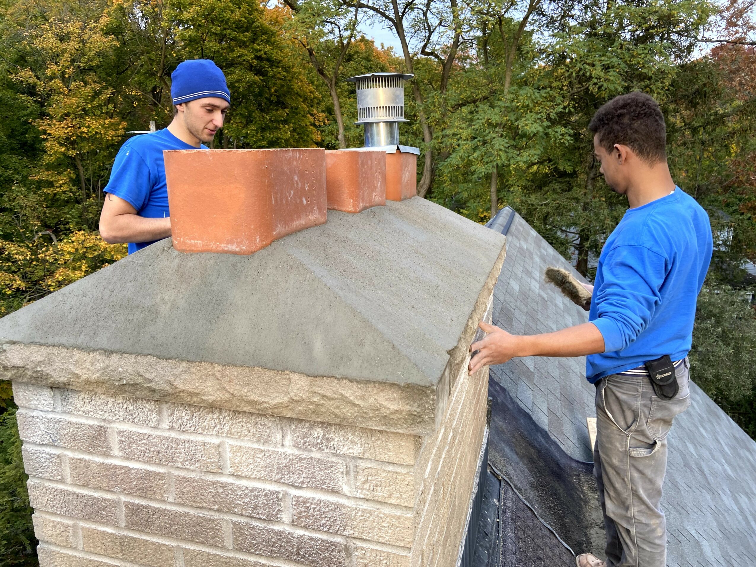 Two boys in blue shirts on a rooftop, one holding a tool, surrounded by trees with autumn foliage.