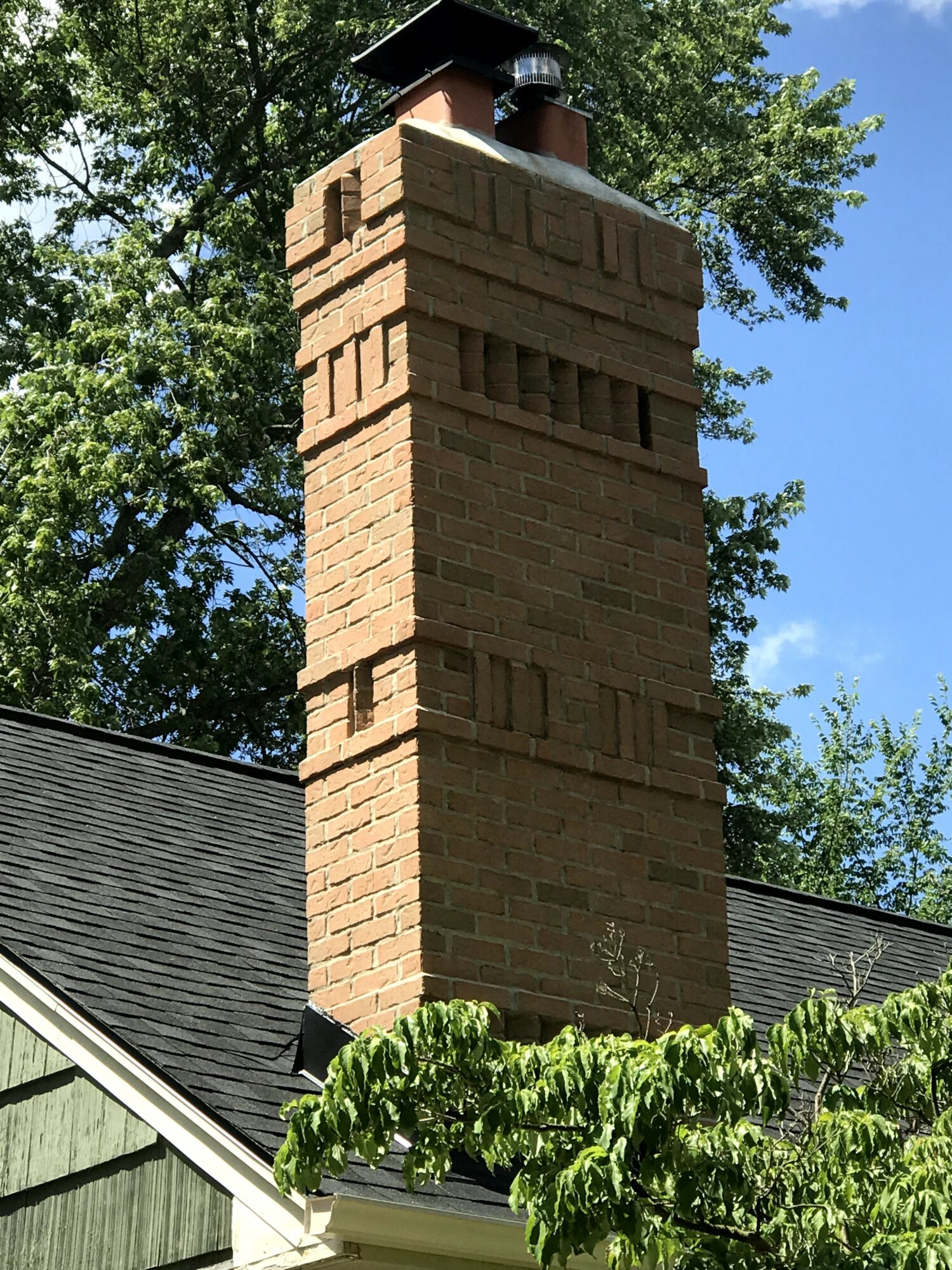 Brick chimney with decorative brickwork on a roof, surrounded by green trees and a clear blue sky.