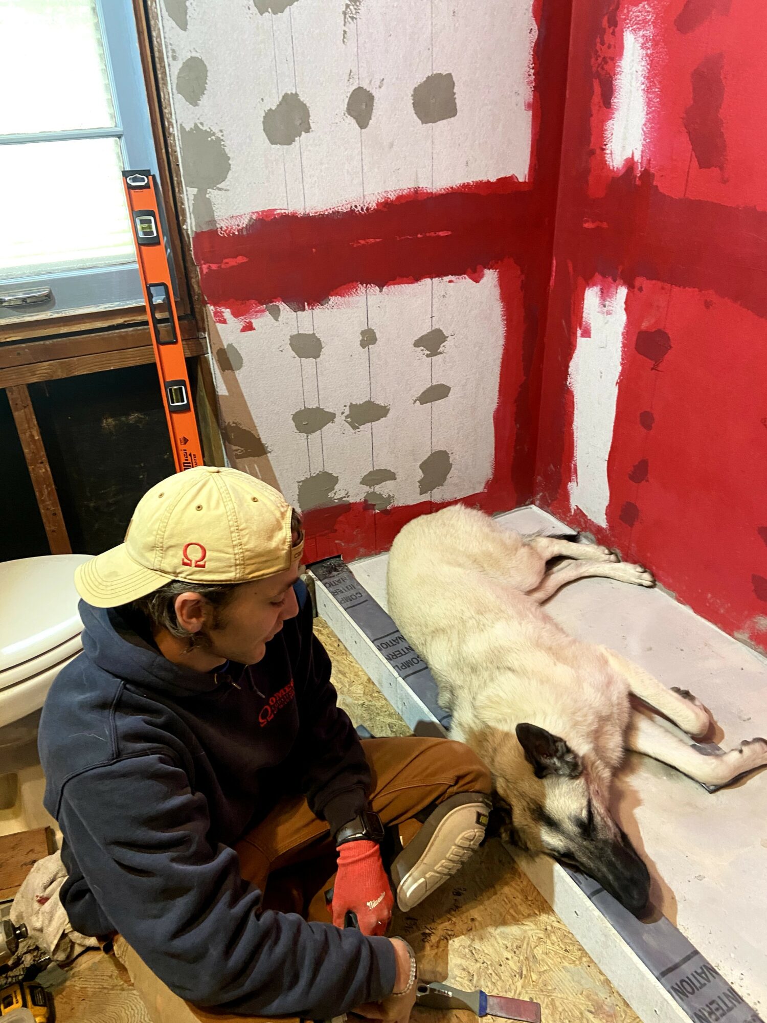 Person in a yellow cap and dark jacket sitting on the floor next to a dog lying down, with a wall under construction in the background.