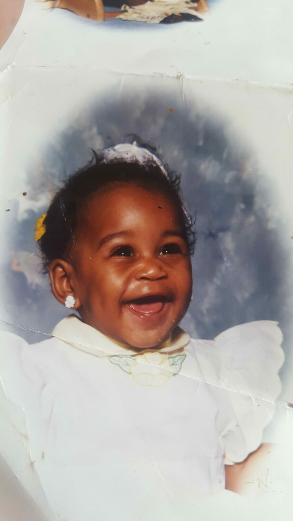 Child with a joyful expression, wearing a white outfit, lying on a surface with a cloudy background.