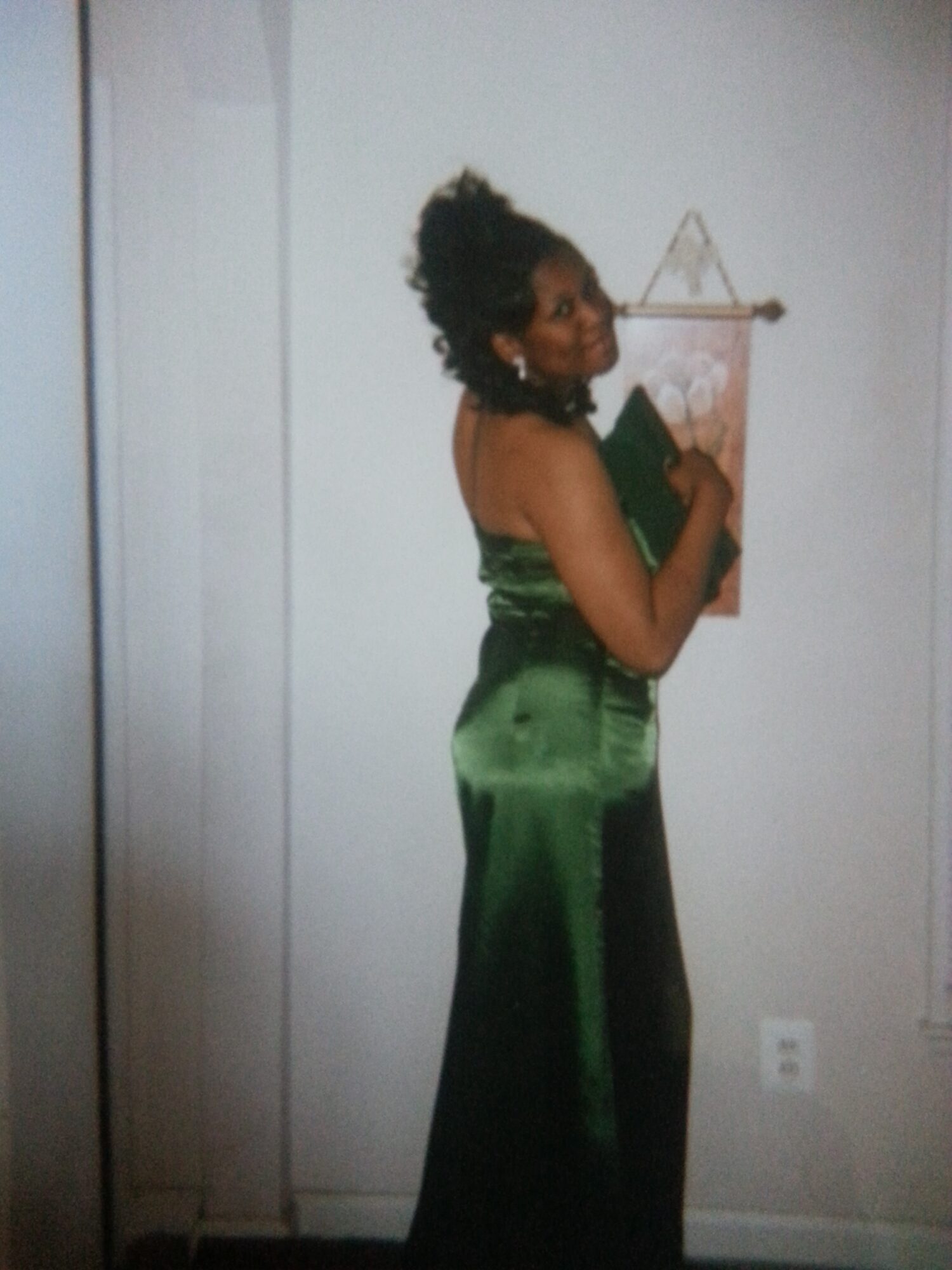 Woman in a green satin dress standing sideways, holding a small object, with a wall and hanging decor behind her.