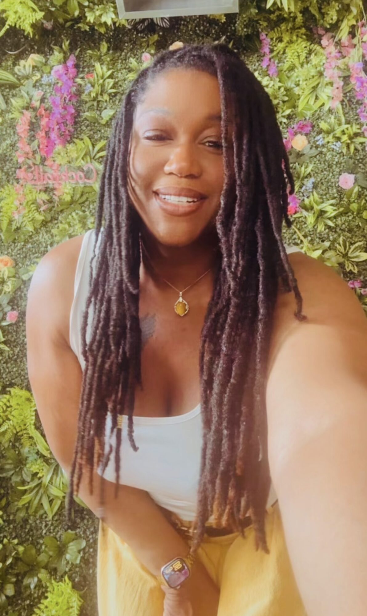 Woman with long dreadlocks smiling outdoors with pink and green flowers in background