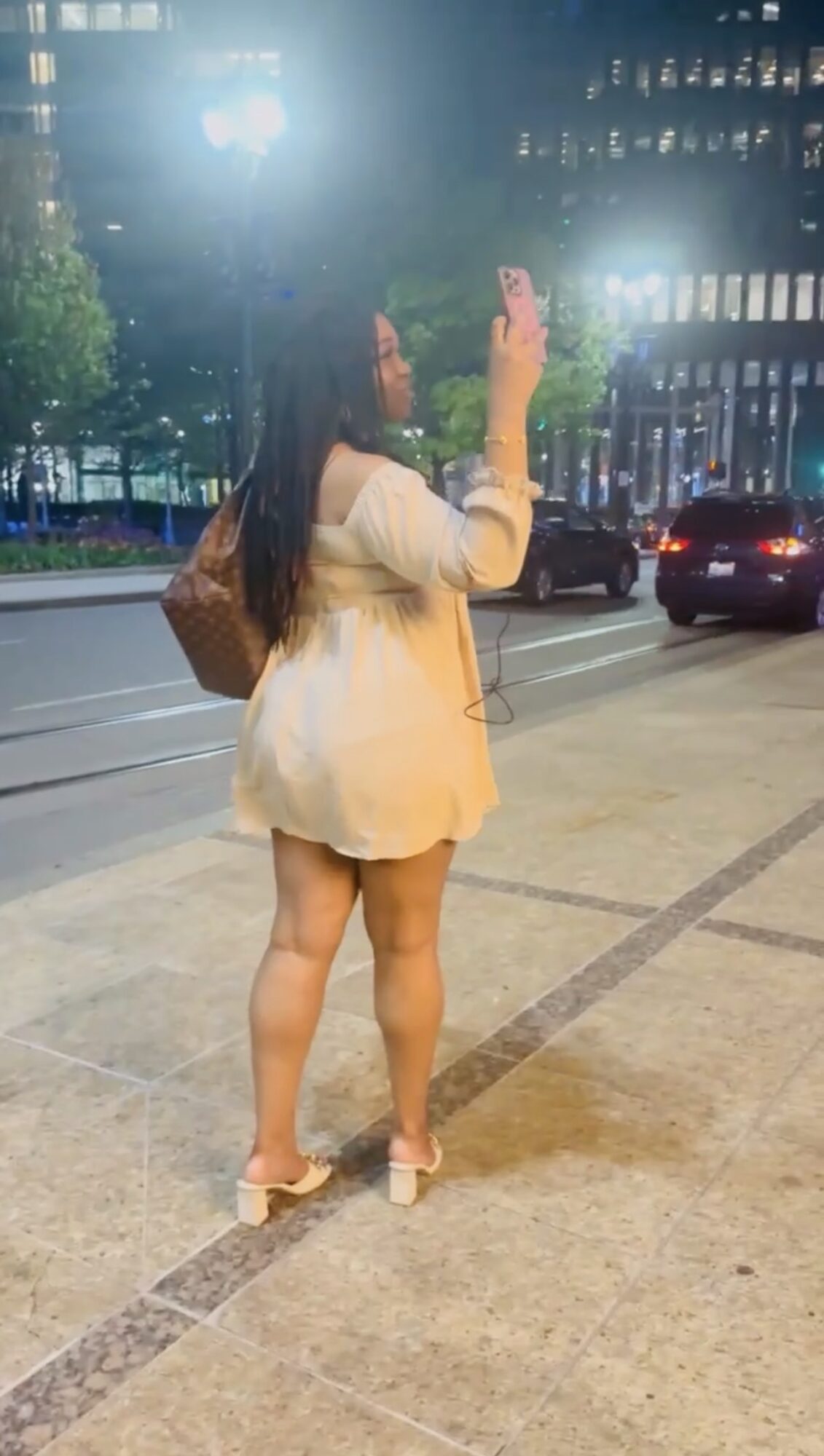 Woman standing on sidewalk taking a photo with her phone at night, city street with cars and buildings in background.