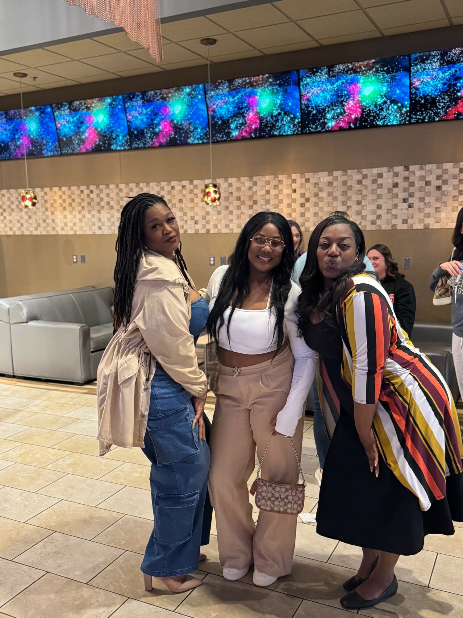 Three women standing together indoors, smiling, with colorful digital display above them.