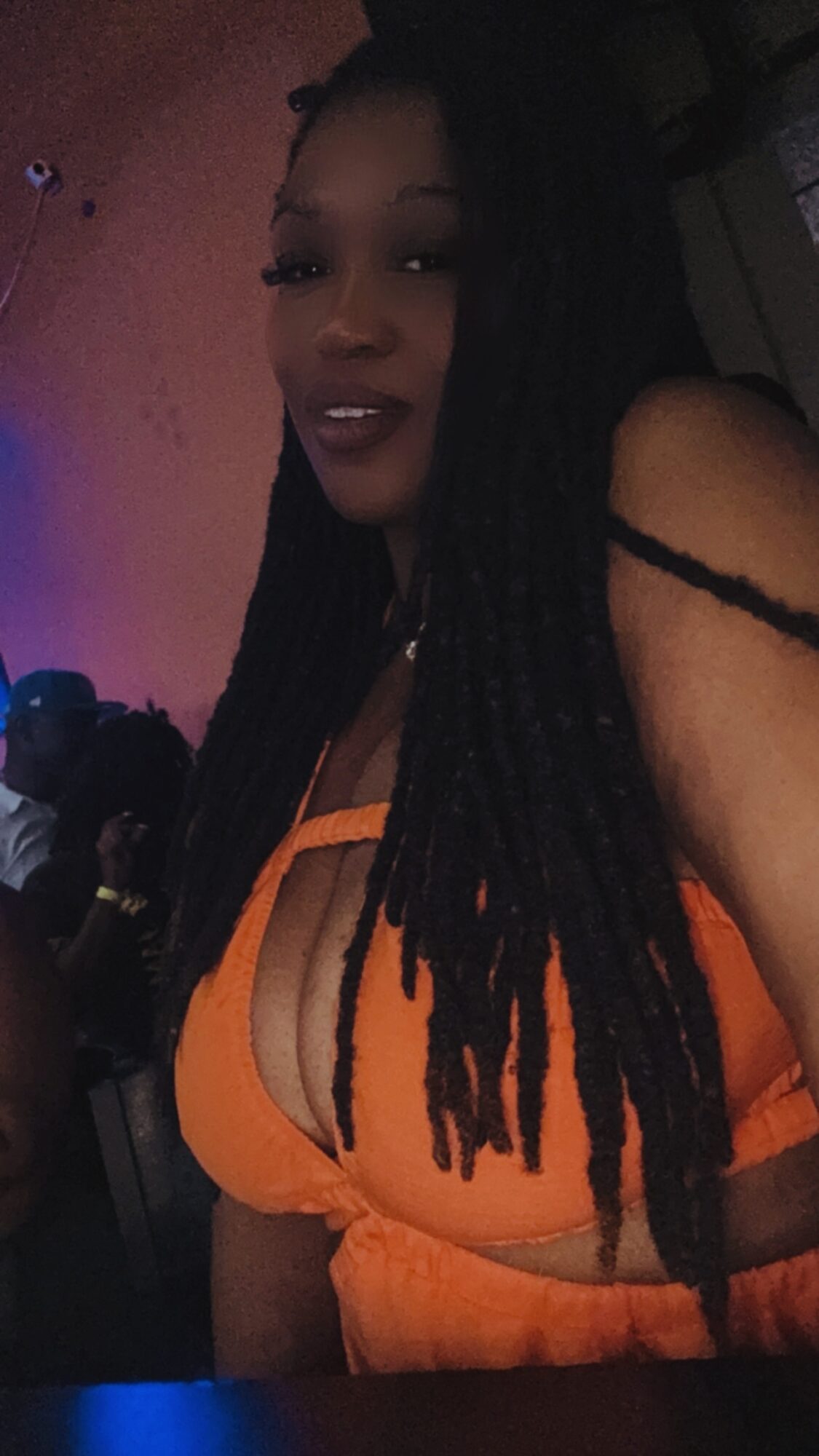 Woman with long braided hair wearing an orange top in a dimly lit environment.