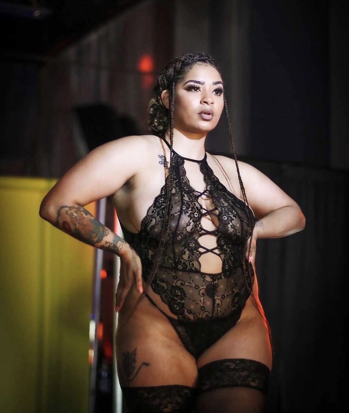 Woman in black lace lingerie with tattoos, standing with hands on hips, in a dimly lit setting.