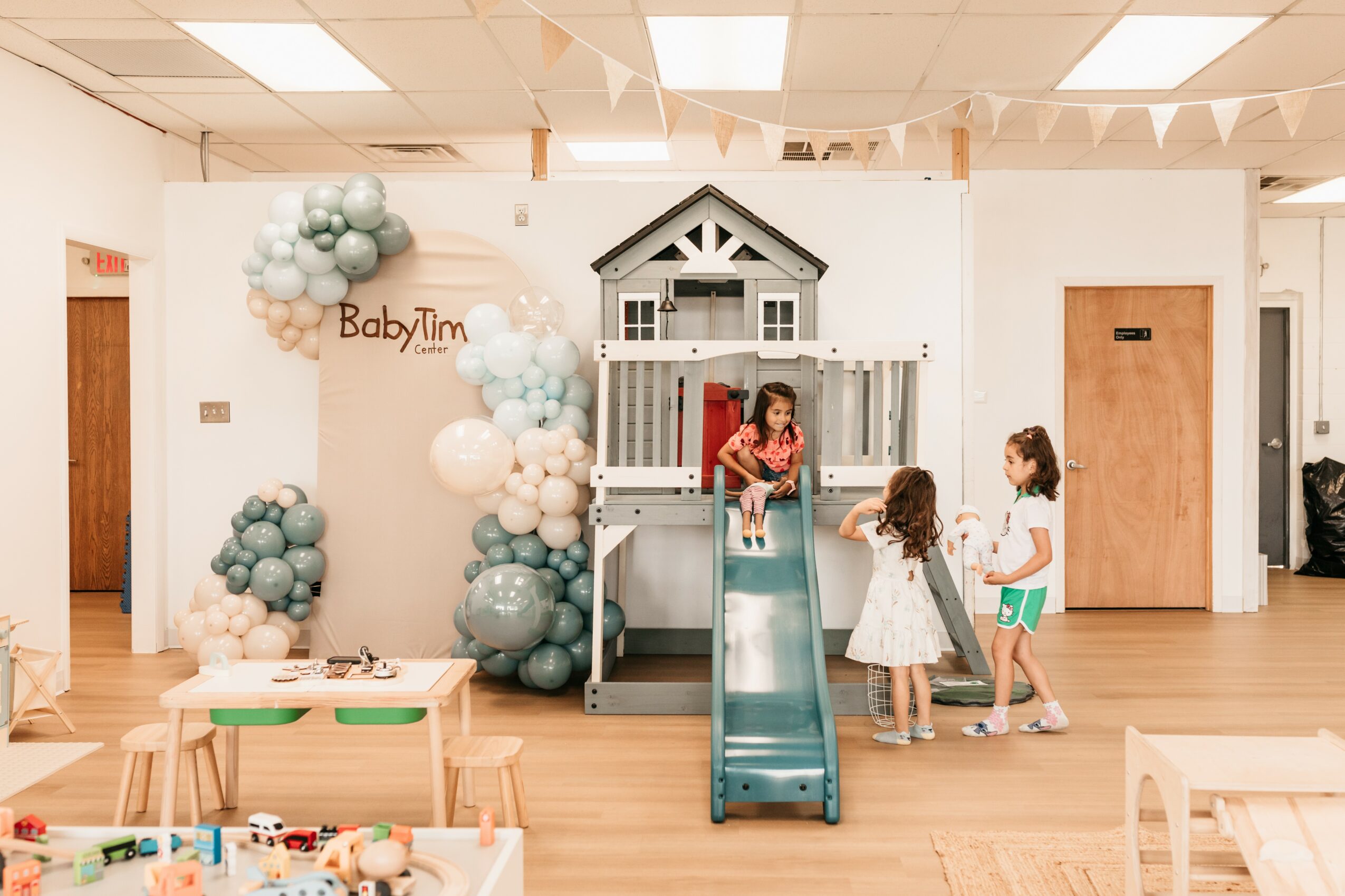 Children playing on a slide at a birthday party with balloon decorations and a small house structure, in a decorated indoor space.