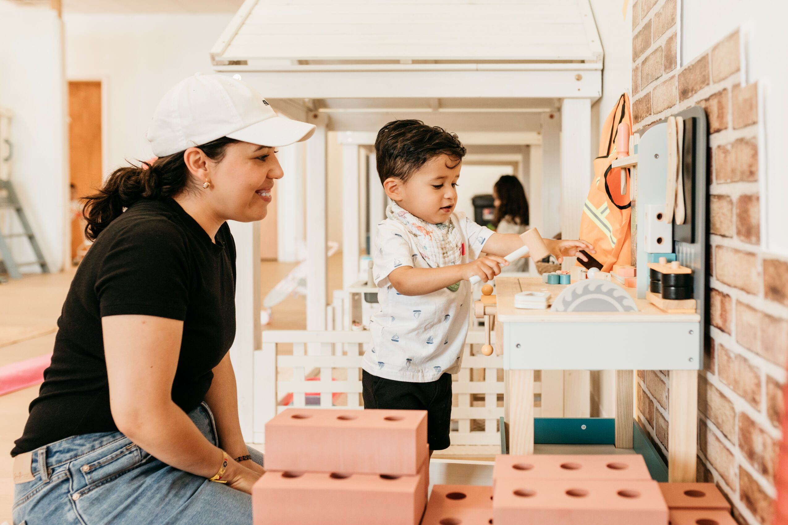 Young boy playing with a toy on a wall-mounted shelf, woman watching, indoor setting with brick wall, children’s play area