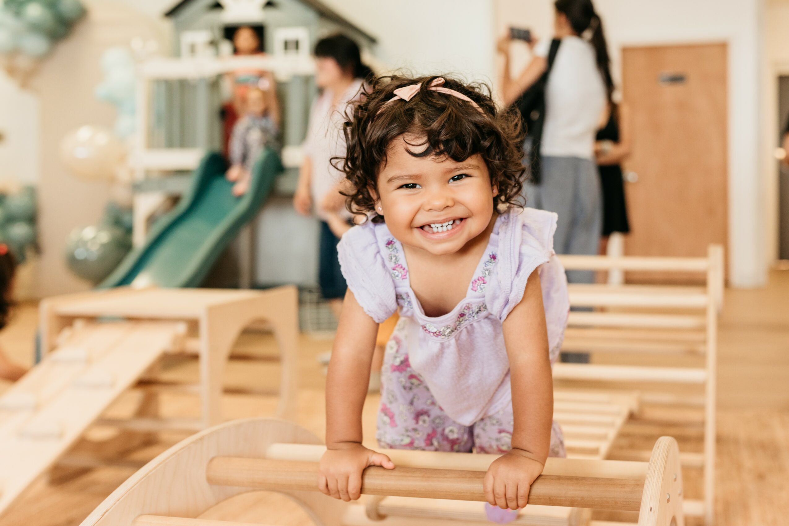 Smiling young girl leaning on a wooden play structure indoors, with people and toys in the background.