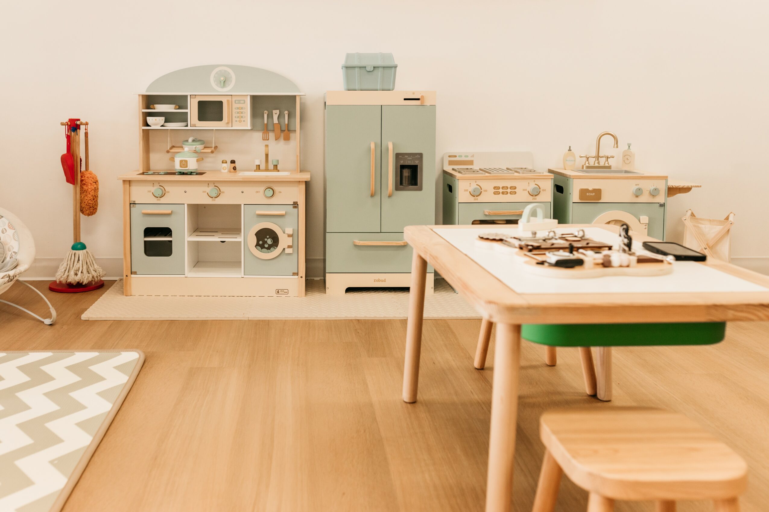 Children's play kitchen set with stove, refrigerator, and sink, and a wooden table with a toy stove on top, in a room with light-colored walls and wooden floor.