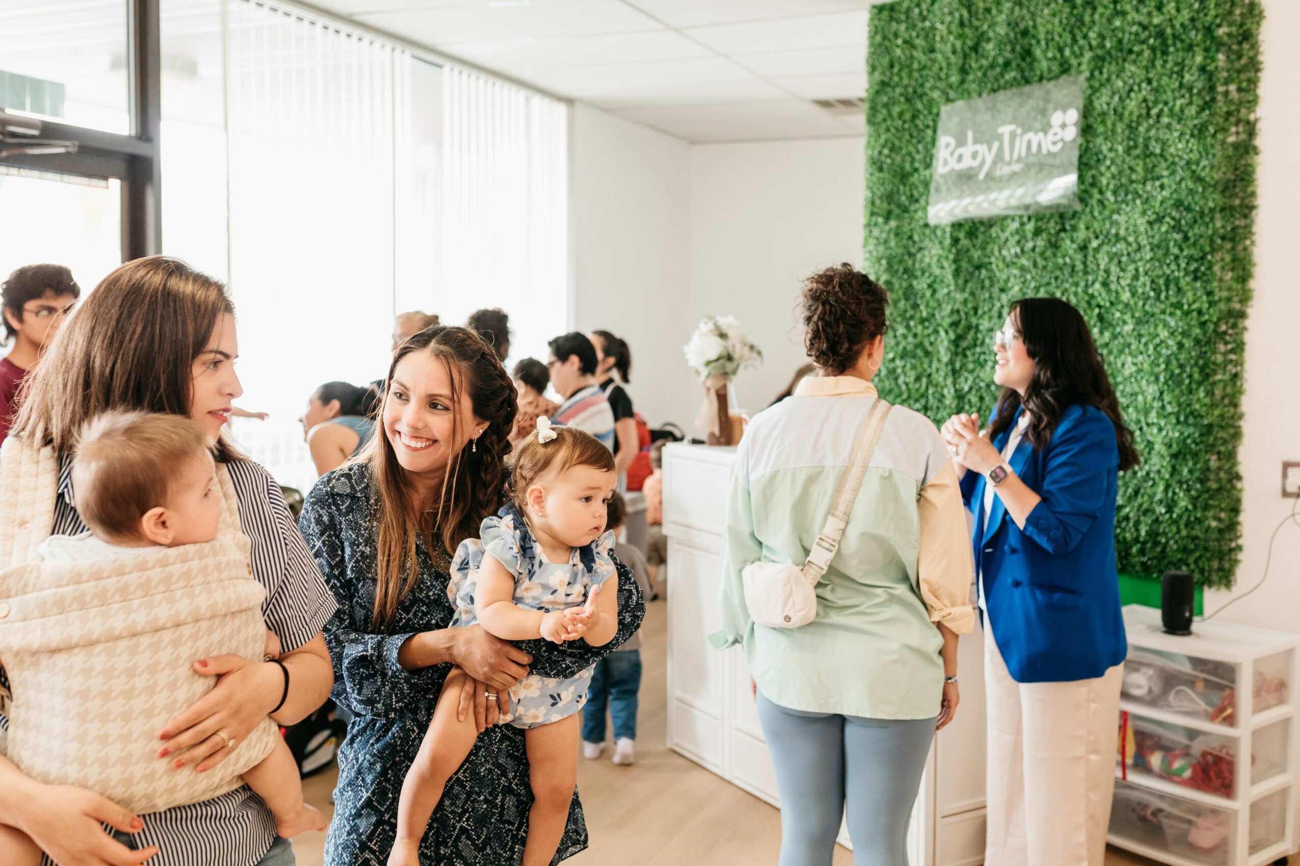 Group of women and children at an indoor event with a green wall and sign reading 'Baby Time'.