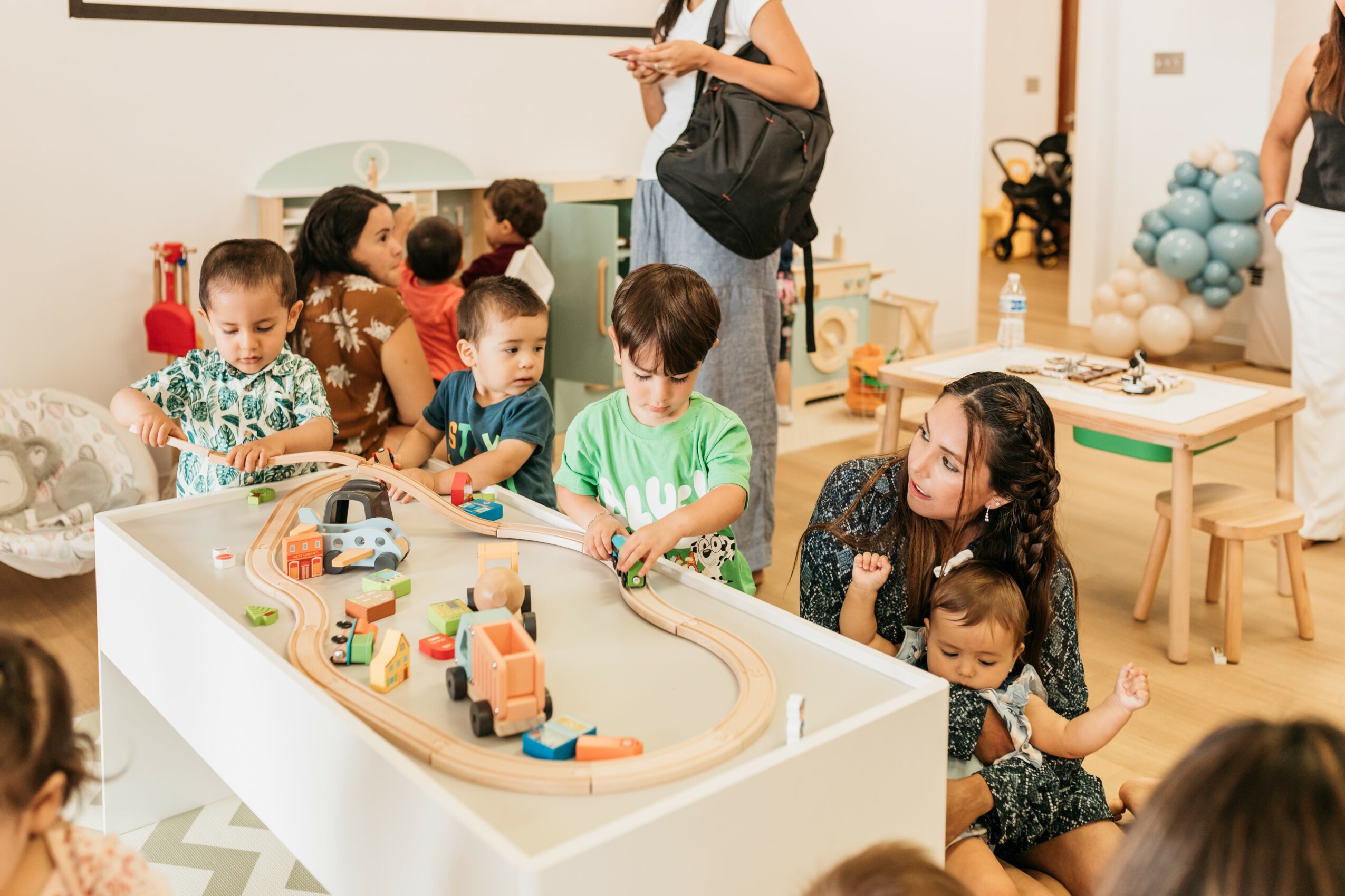 Children and adults play with a train set on a white table in a room with balloons and toys, some sitting and some standing.