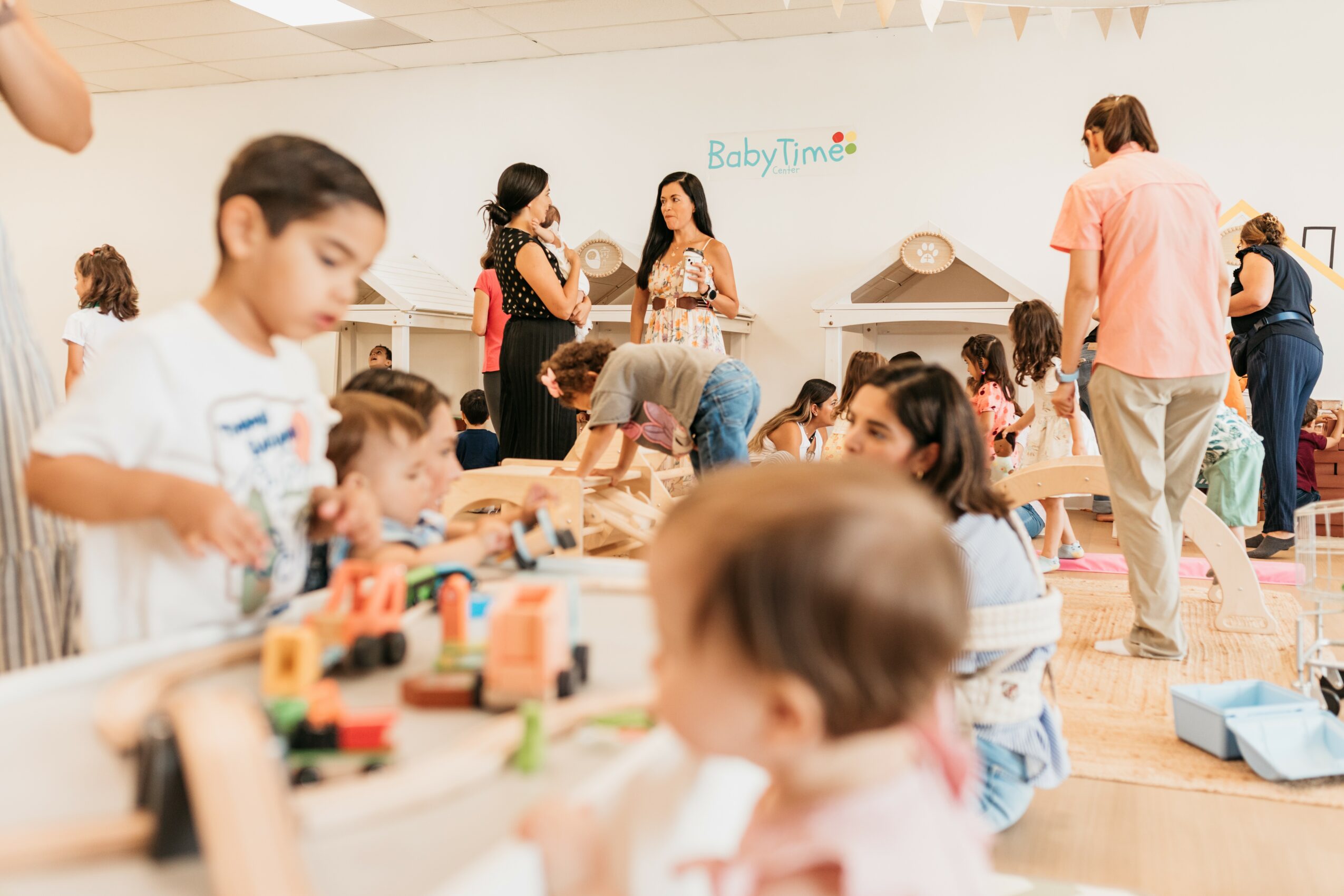 Children and adults in a playroom with toys and activities, some sitting on the floor, others standing or walking, with a sign reading 'BabyTime' on the wall.