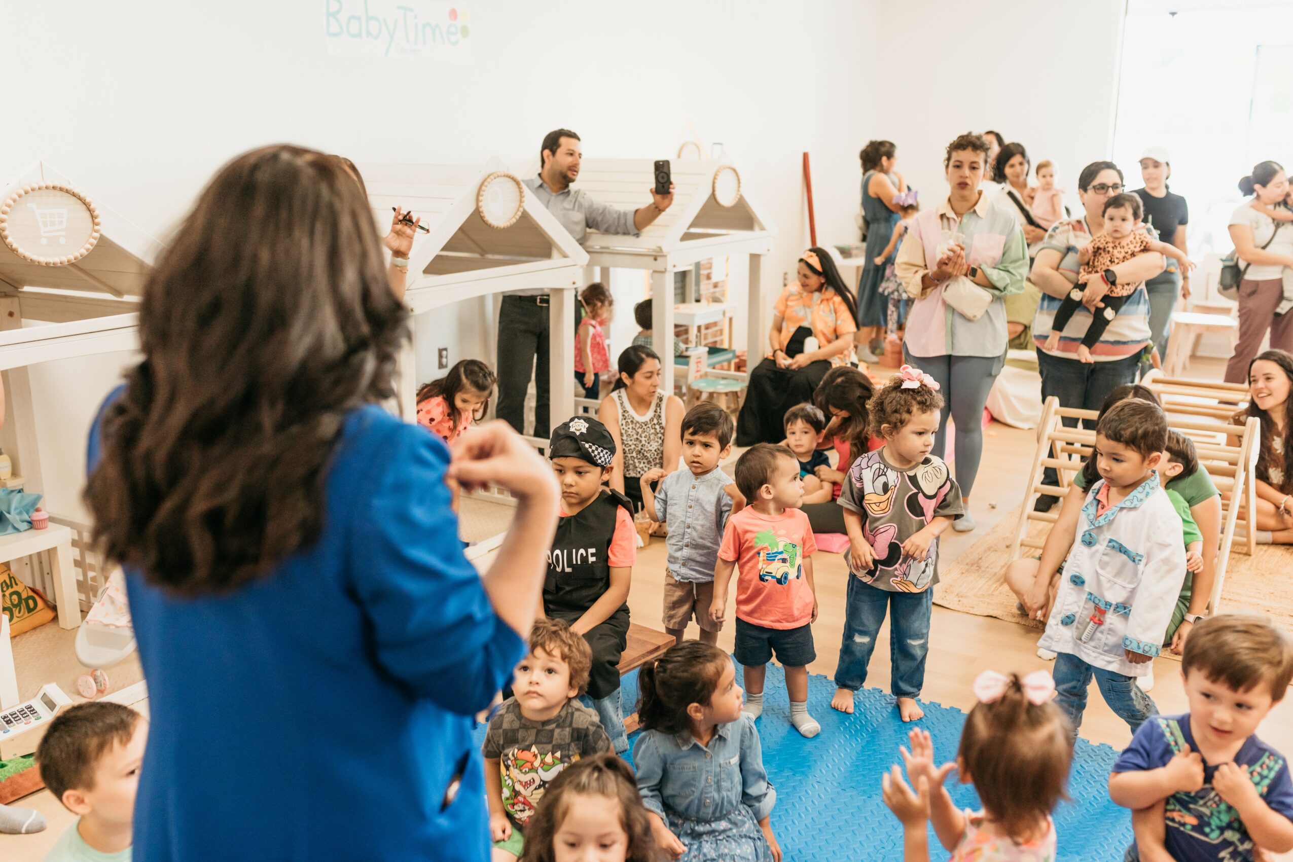 Group of children and adults in a classroom, with a woman in blue in foreground, some children sitting and others standing.