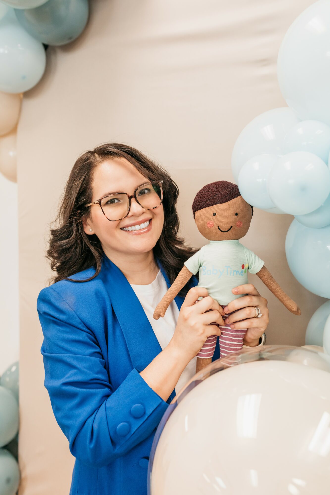 Woman with glasses and a blue blazer holding a doll, smiling, surrounded by light blue and white balloons.