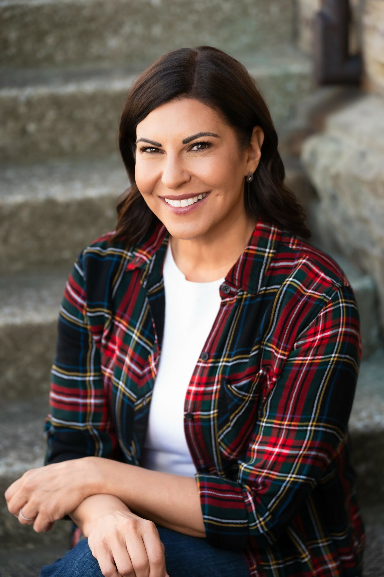 Smiling woman with dark hair, wearing a plaid shirt, sitting outdoors on stone steps.