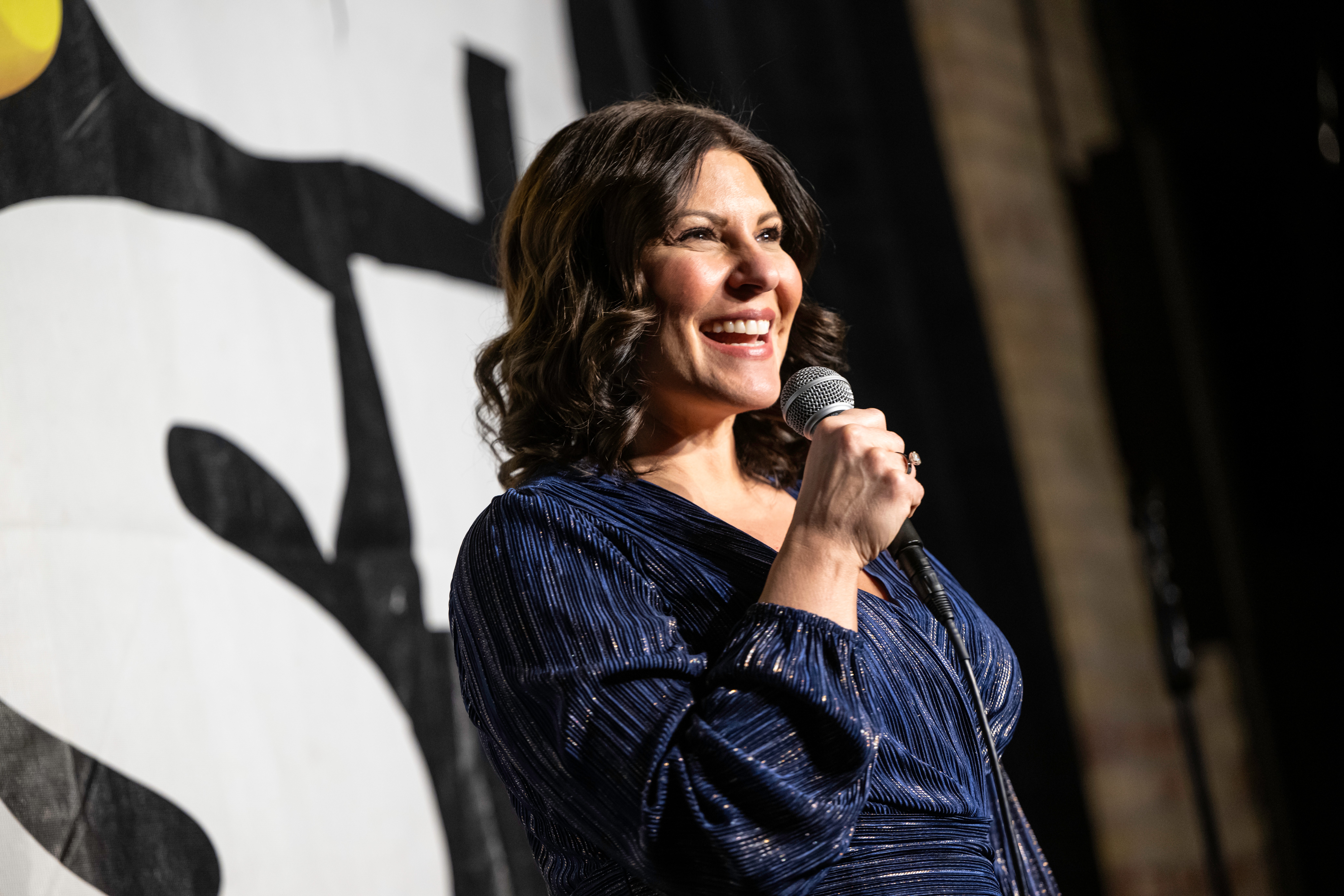 Woman smiling and speaking into a microphone on stage, with a black and white background behind her.