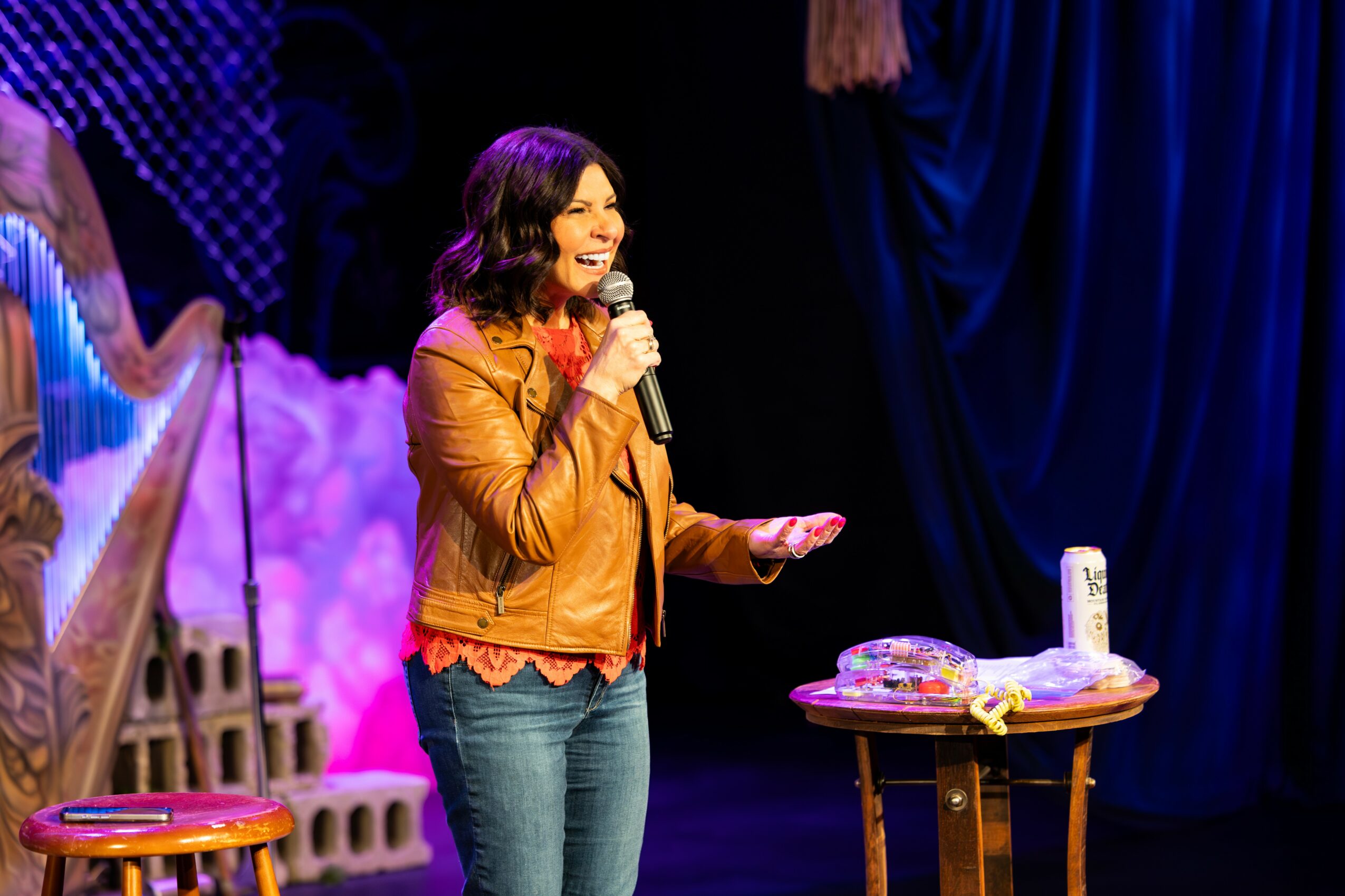 Woman holding microphone on stage with table and props, dark blue curtain background, smiling, performing.