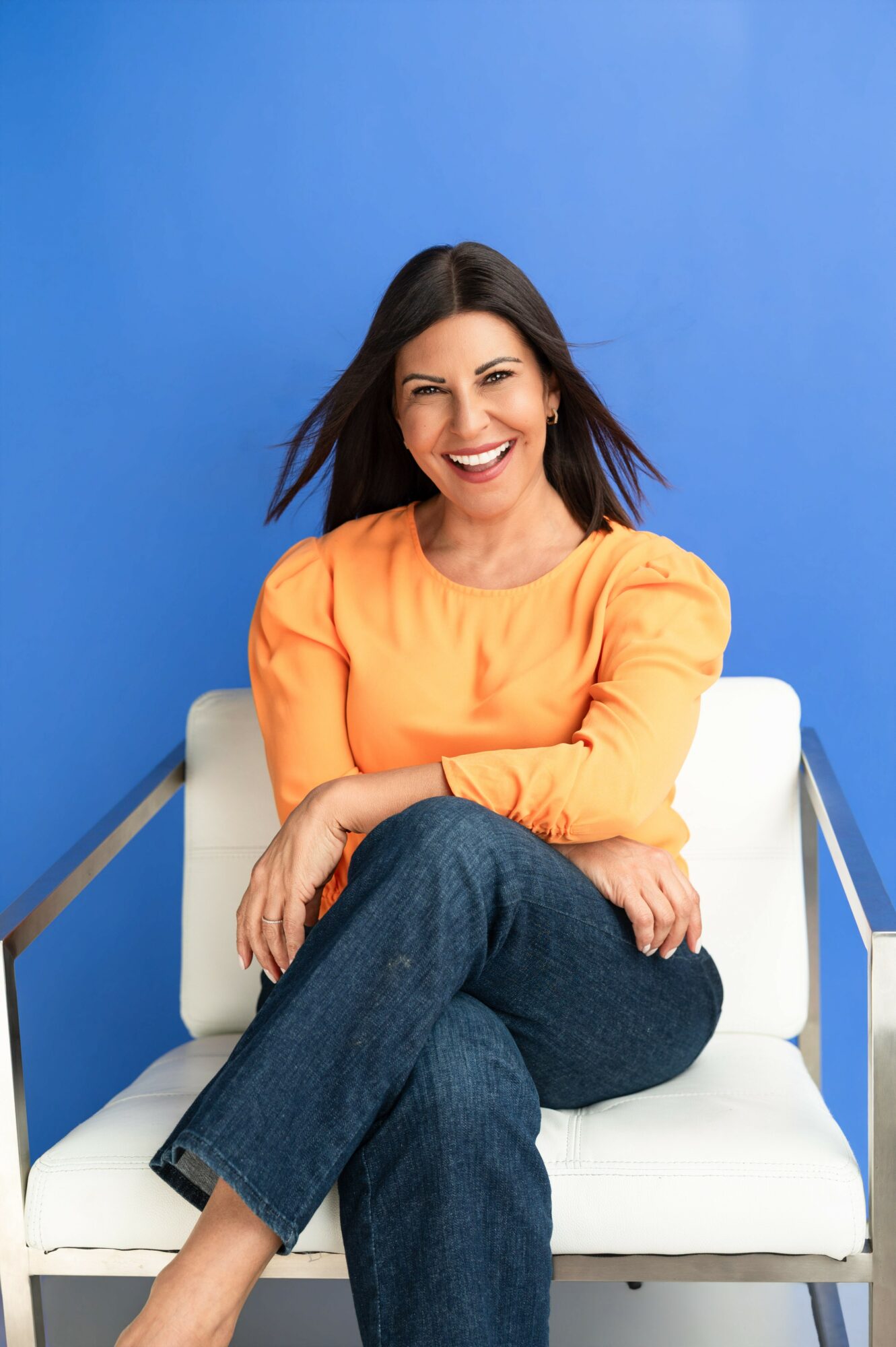 Woman with dark hair smiling, wearing an orange top and jeans, sitting on a white chair against a blue background.