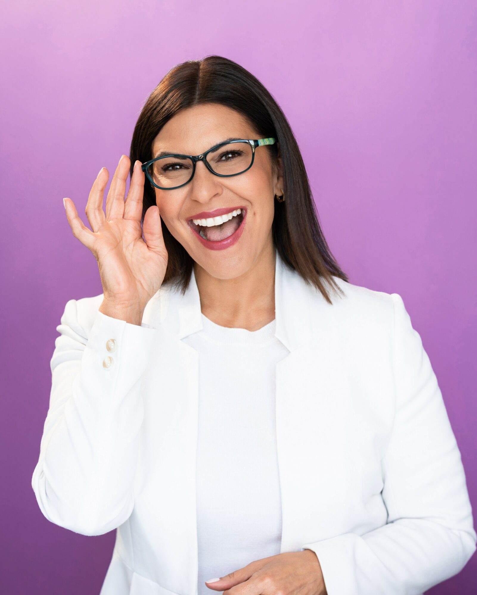 Woman with glasses smiling and holding her hand near her face against a purple background.