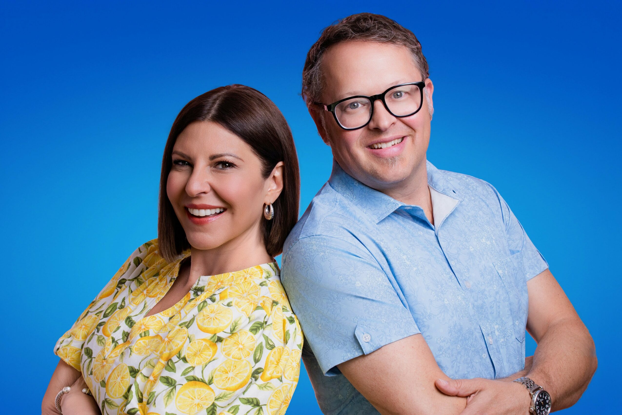 A smiling woman and man stand side by side against a blue background, both with arms crossed, looking at the camera.
