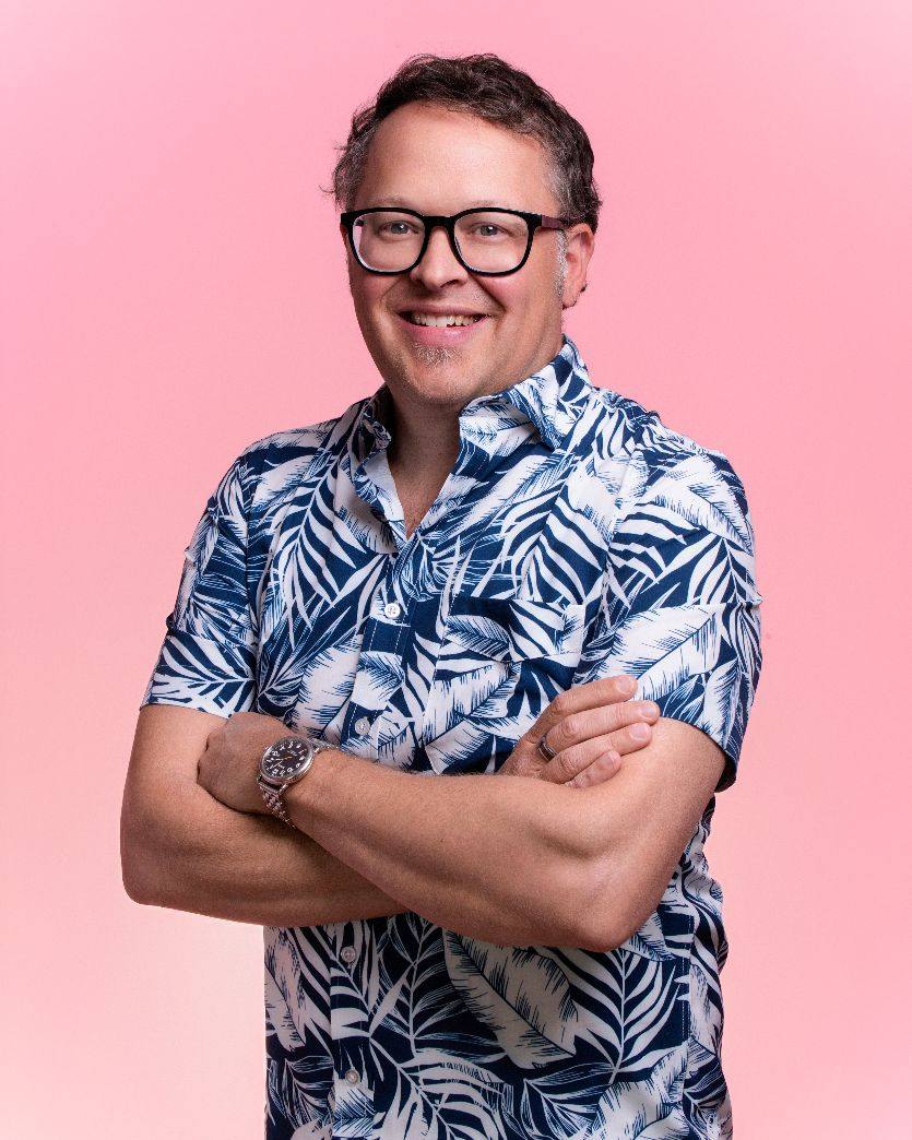 Man with glasses smiling, arms crossed, wearing a blue and white tropical shirt, against a pink background.