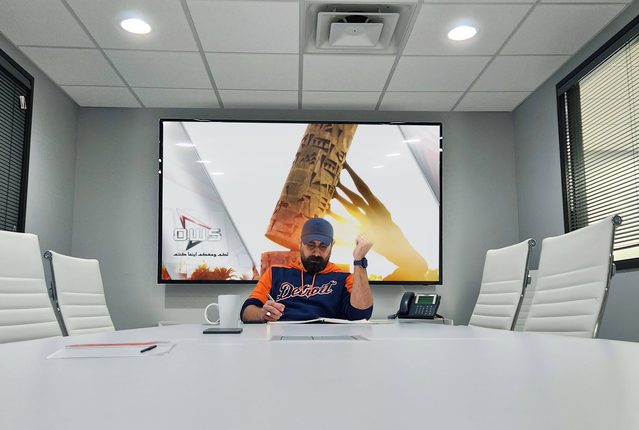 Person sitting at a conference table with a large screen behind, showing an image of a hand holding a model of the Eiffel Tower.
