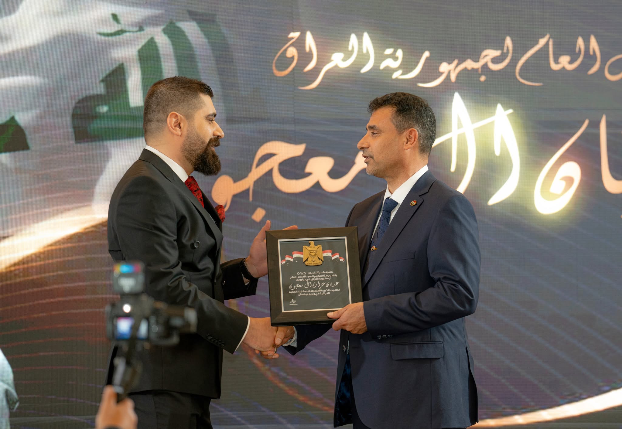 Two men in suits exchange a framed certificate on stage with Arabic script in the background.