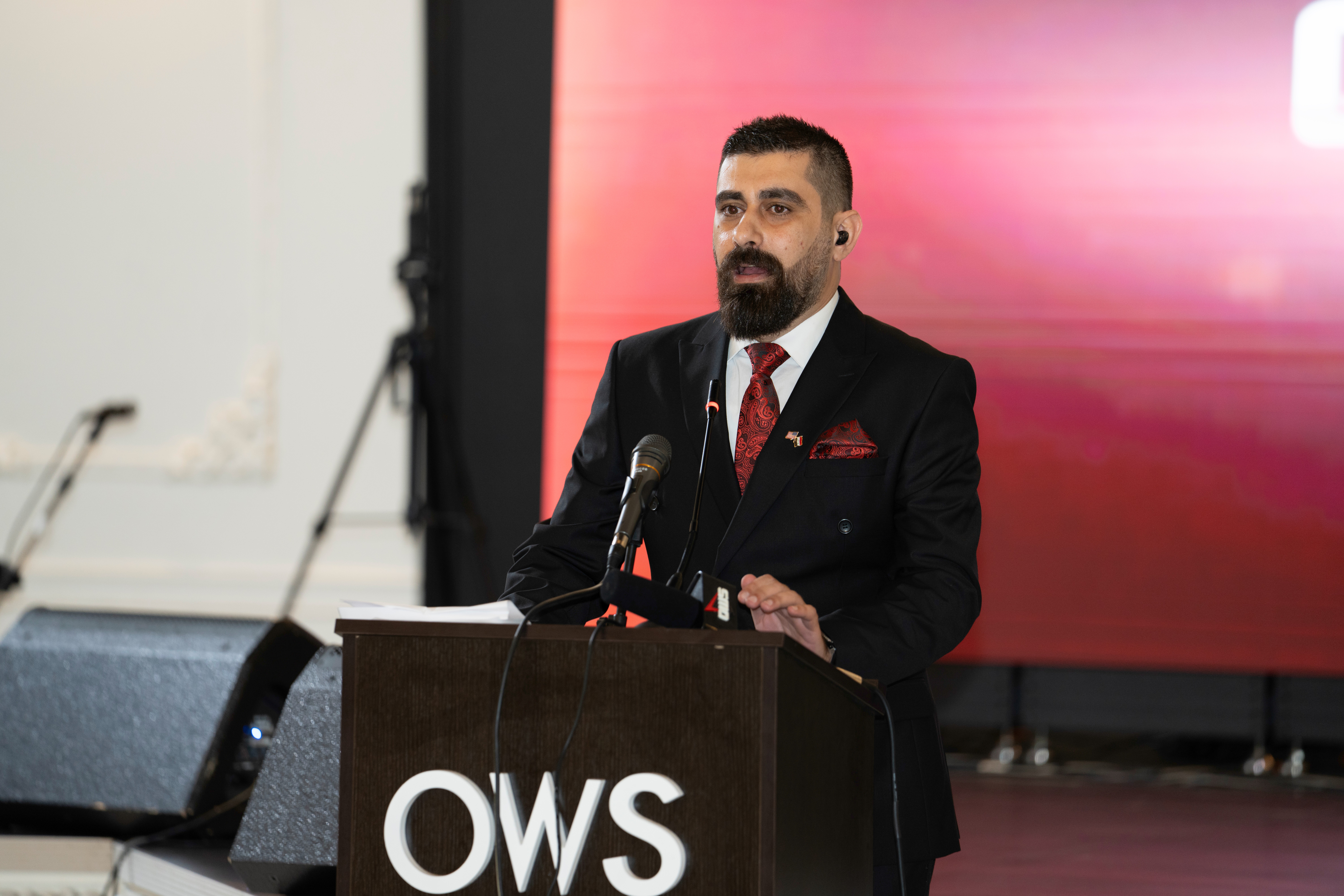 Man in suit speaking at a podium with a microphone, behind a red and pink gradient screen, in a conference setting.