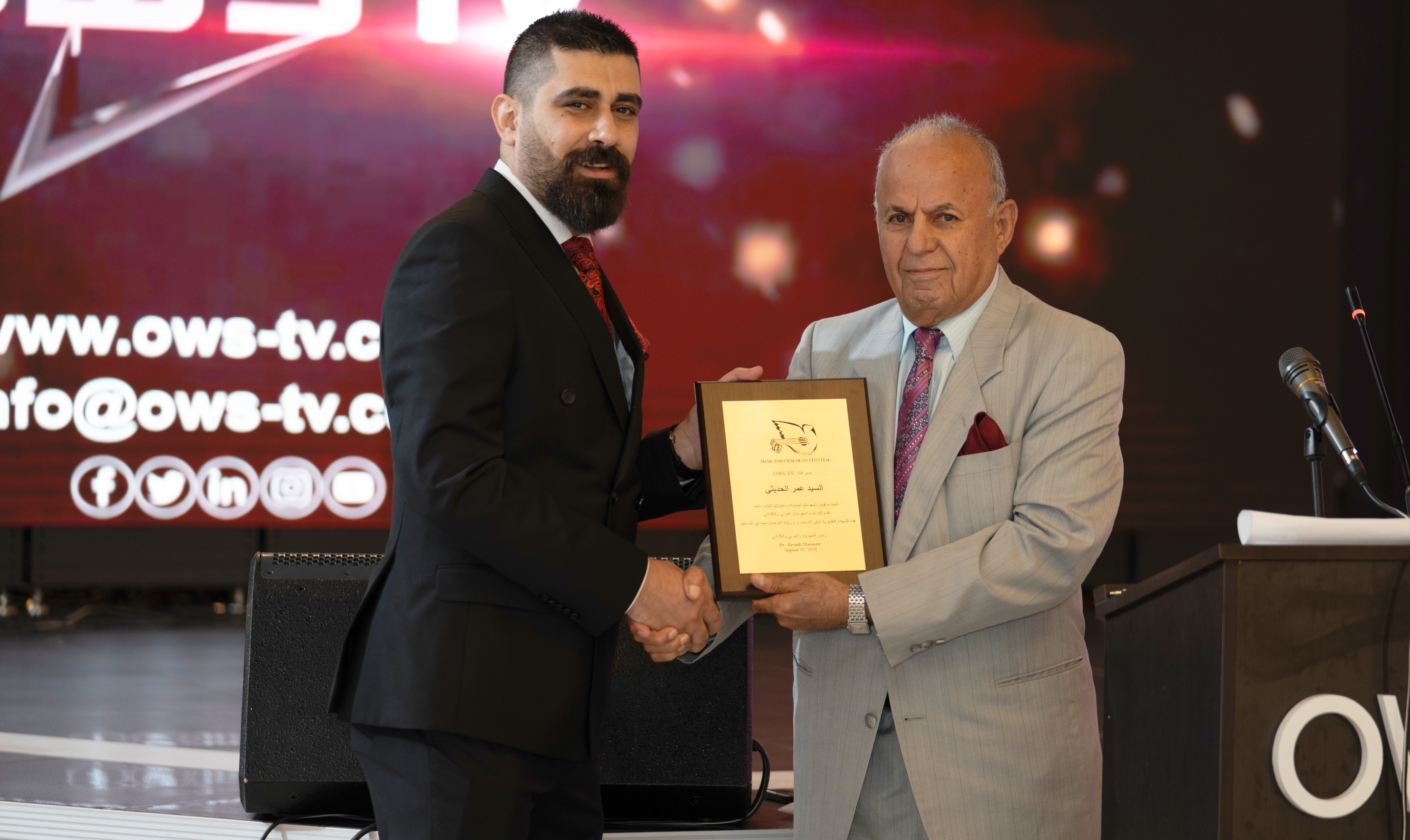 Two men in suits shaking hands, one holding a framed certificate, in a formal event setting with a screen in the background.