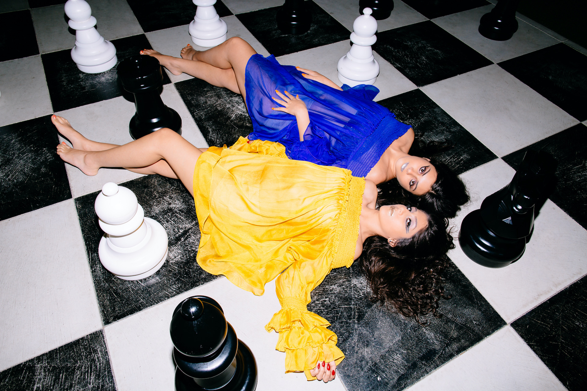 Two women in colorful dresses lie on a black and white checkered floor among large chess pieces.