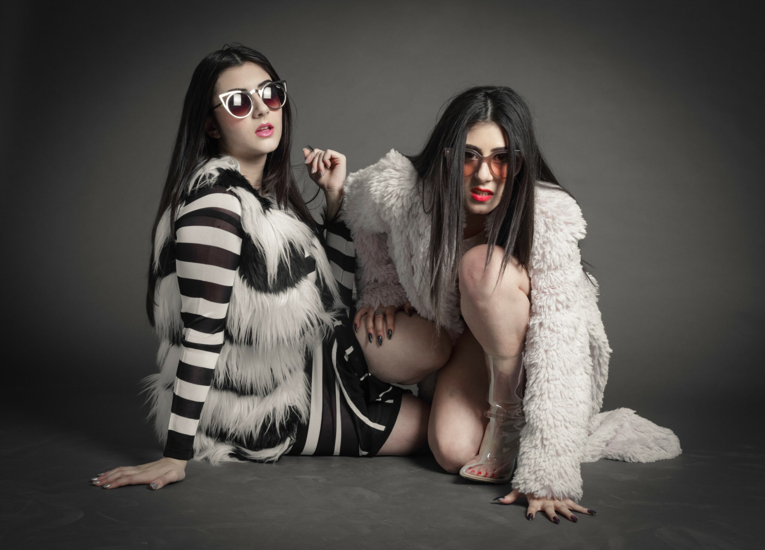 Two women with long dark hair and sunglasses pose on a dark background, one wearing a striped fur coat and the other a fluffy coat, both in provocative poses.