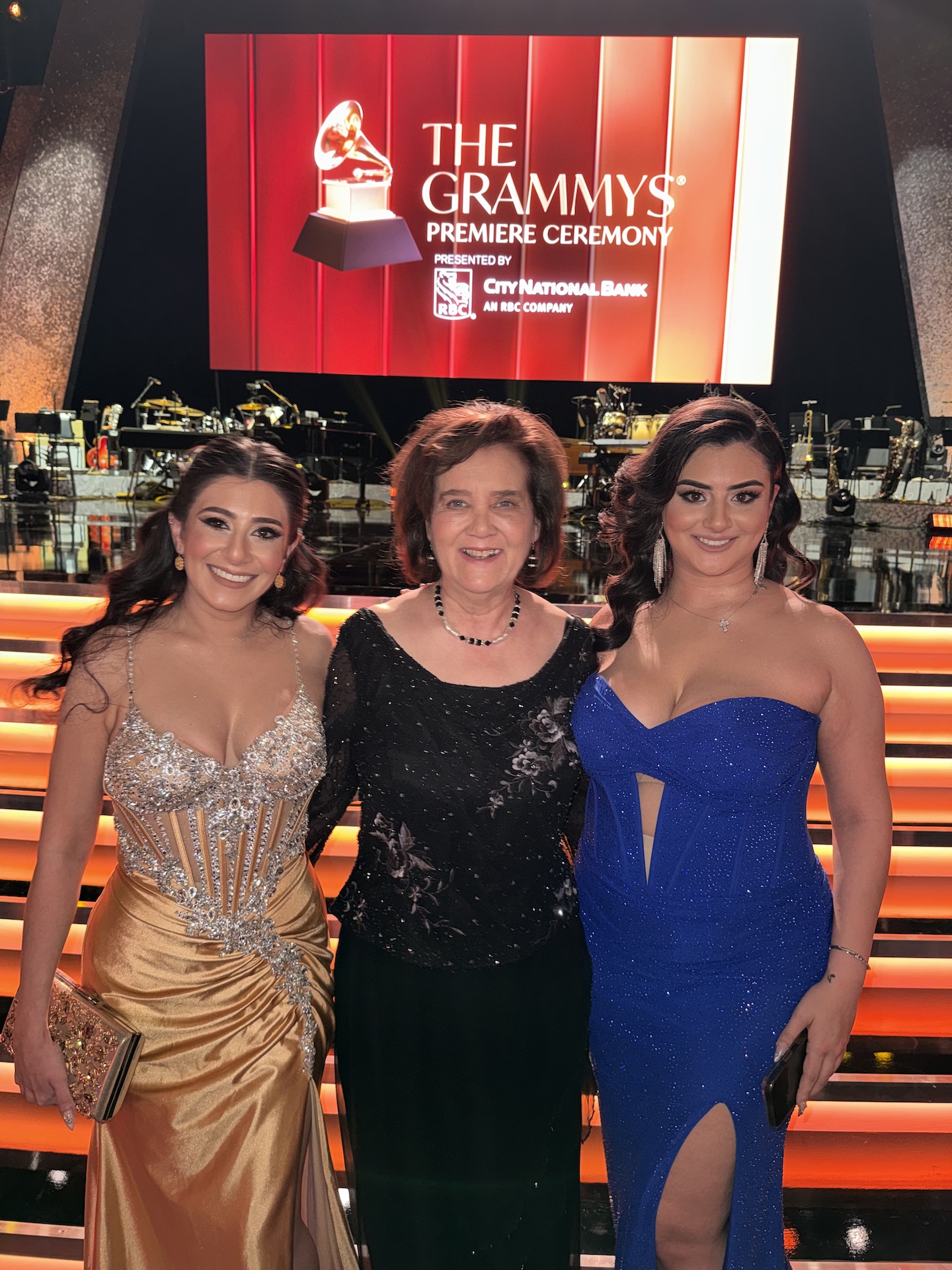 Three women standing together at an event, with a large screen behind them displaying 'The Grammy's Premiere Ceremony' logo.