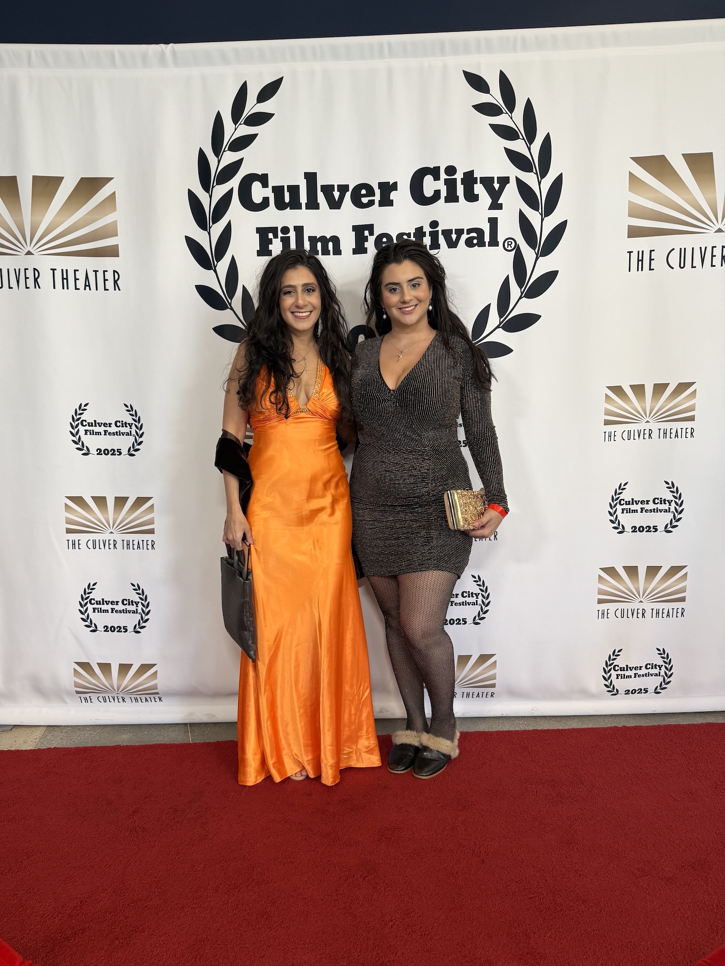 Two women standing on a red carpet in front of a backdrop with festival logos, smiling, one in an orange dress, the other in a black dress.