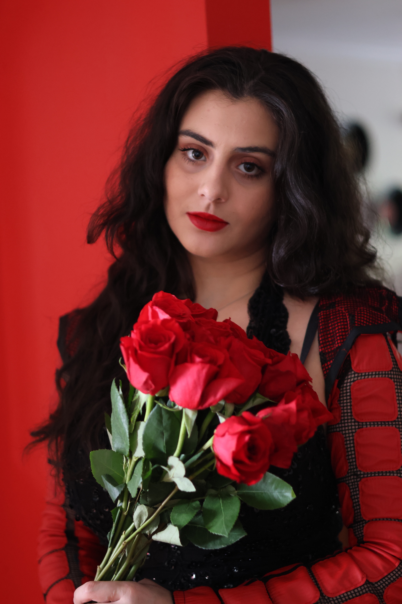 Woman with long dark hair holding a bouquet of red roses, wearing a red and black dress, standing against a red background.