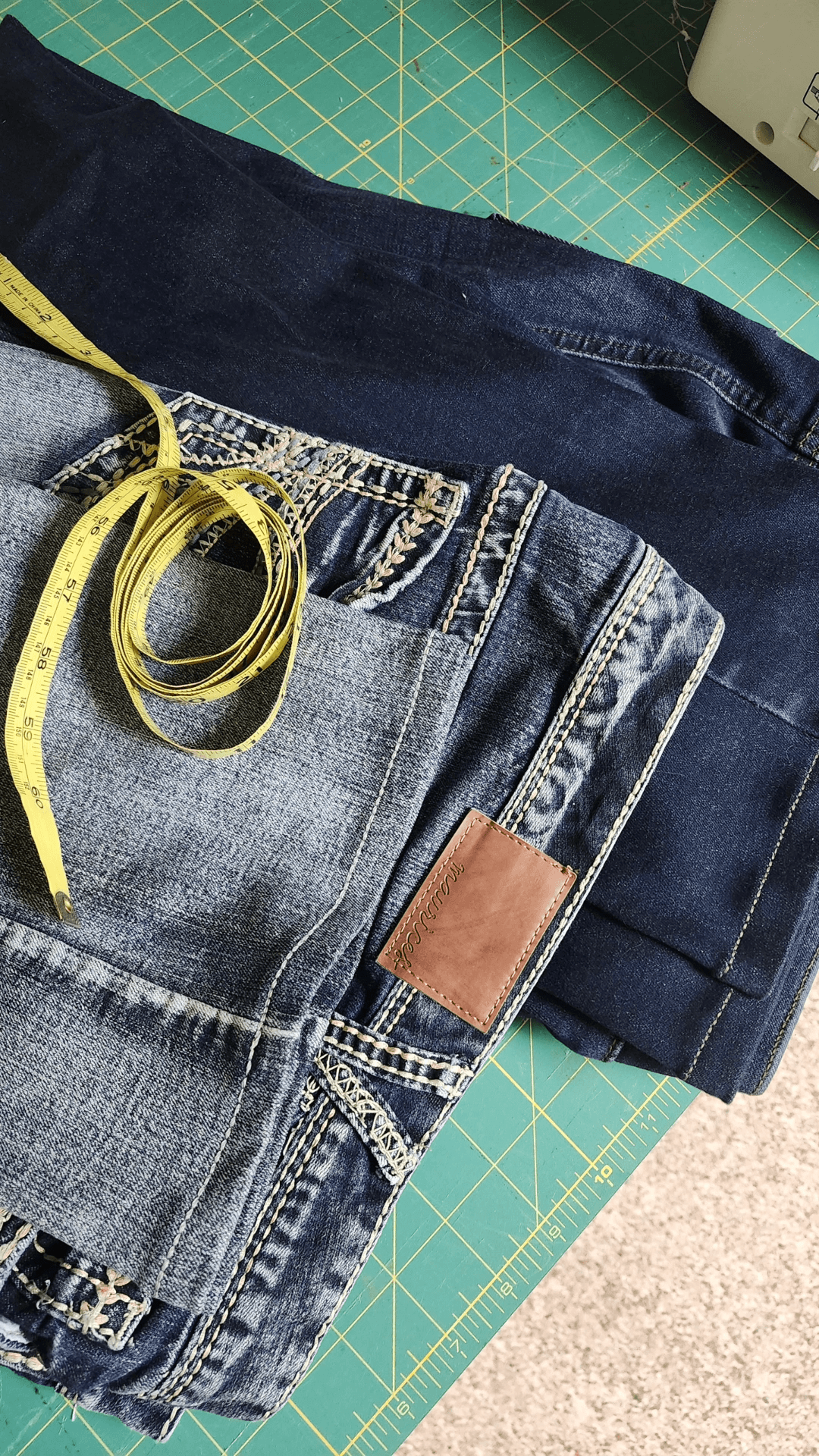 Two pairs of jeans, one dark and one light, with a yellow measuring tape on top, on a green cutting mat.